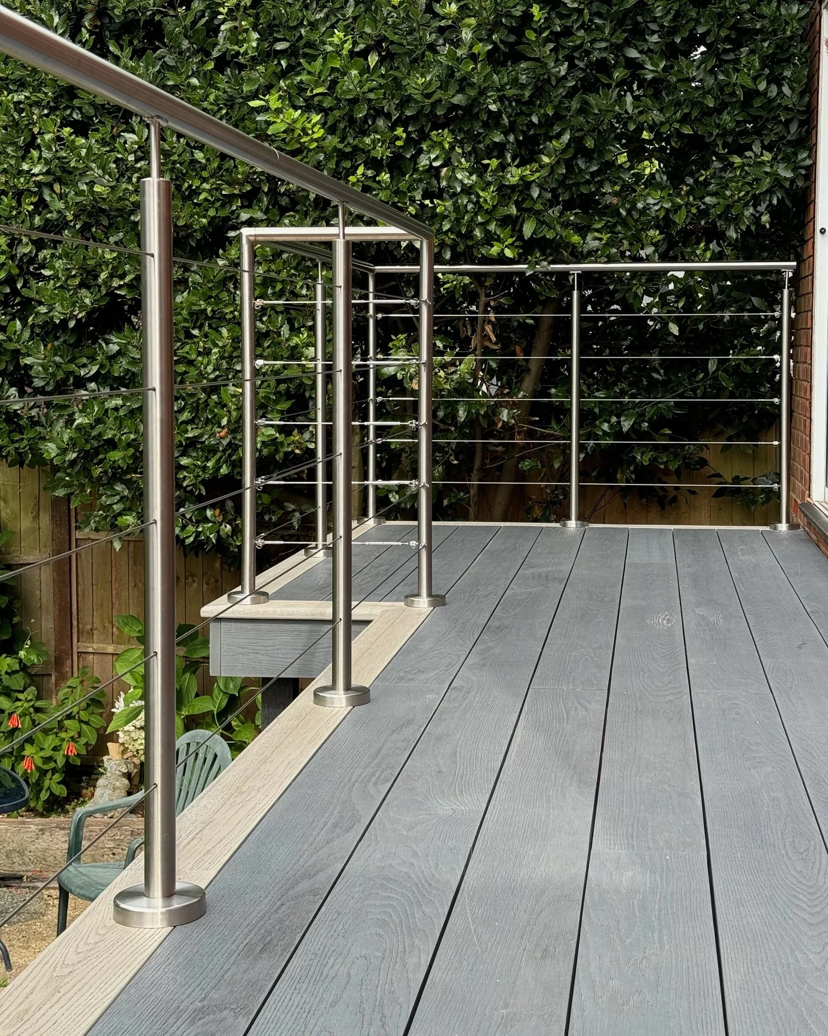 One of Esme's decking and balustrading projects which was to install a new outdoor deck with gray millboard composite flooring, stainless steel railing, and a background of lush green hedges.