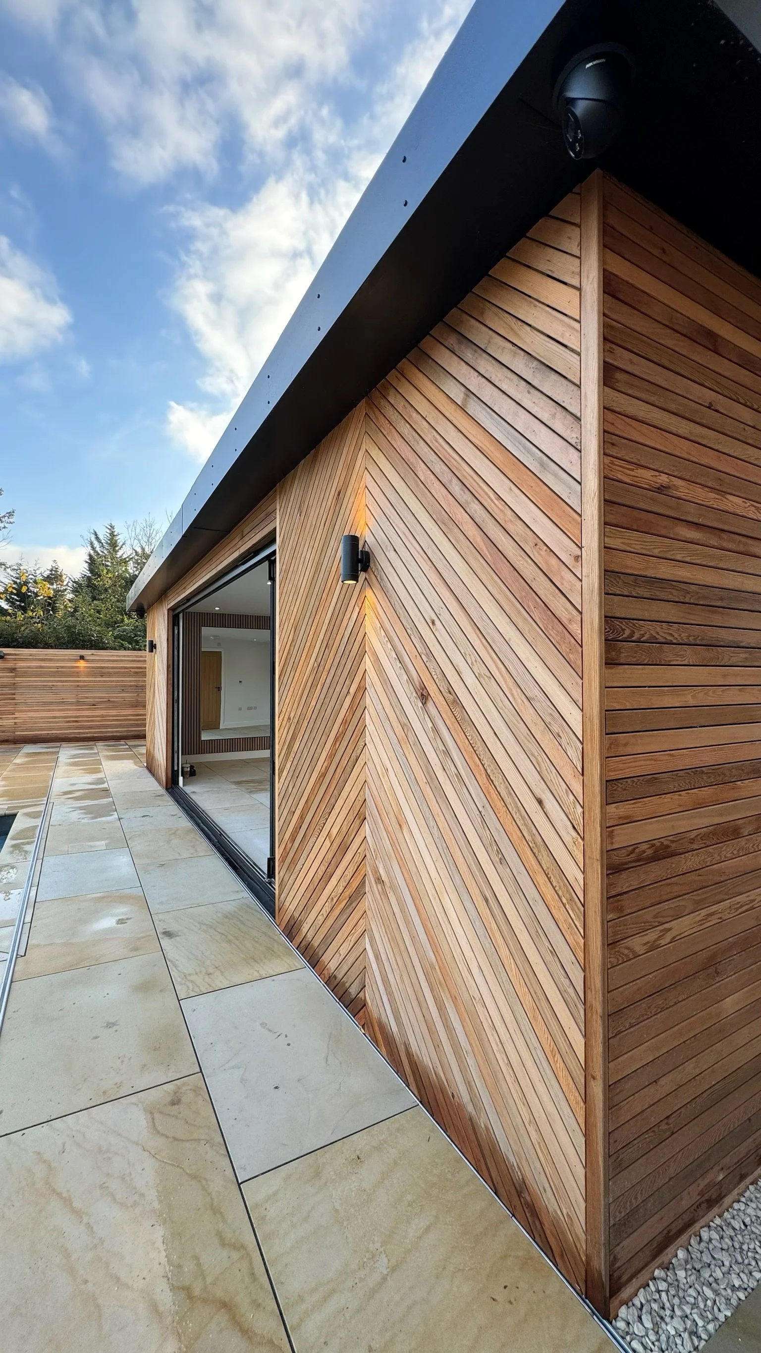 Modern house with wooden exterior and tile patio, sliding glass door open to an interior room, under a partly cloudy sky.