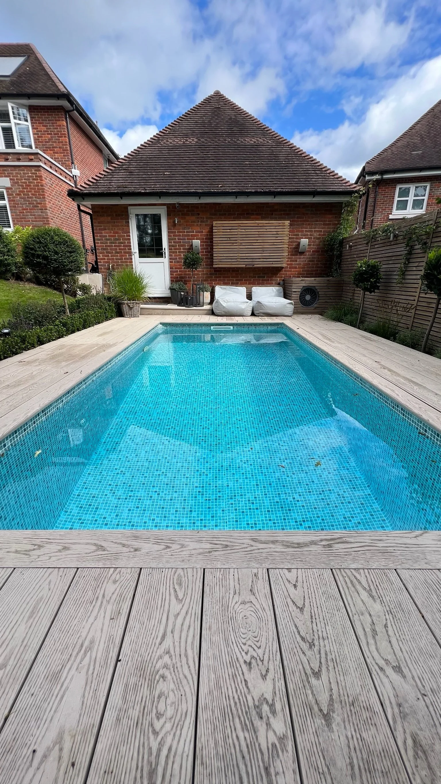Rectangular swimming pool with blue mosaic tiles, surrounded by light-colored wooden deck, facing a brick house with a sloped roof, outdoor seating, and plants under a partly cloudy sky.