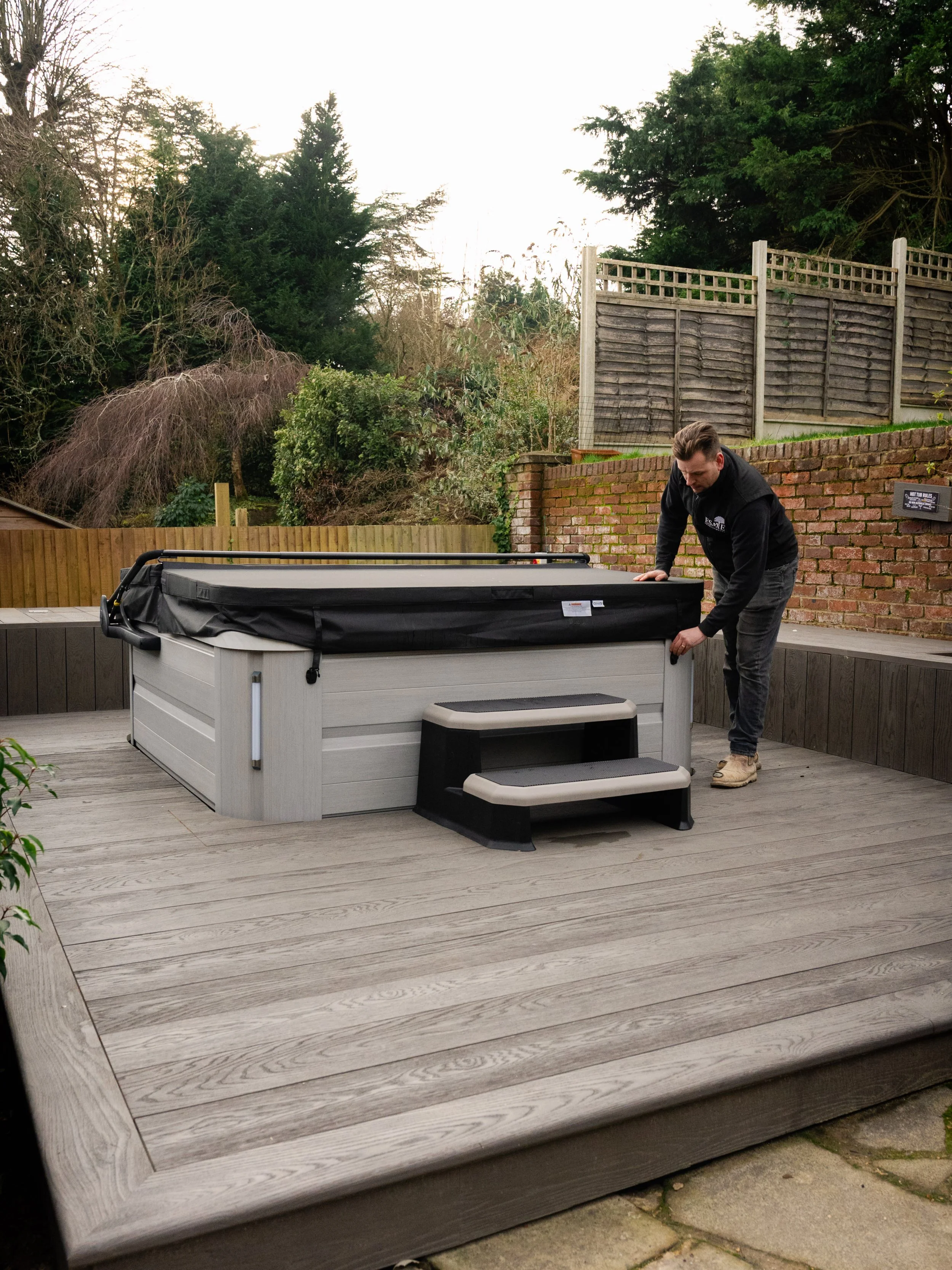 A man setting up a hot tub on a wooden deck in a backyard with trees and fencing.