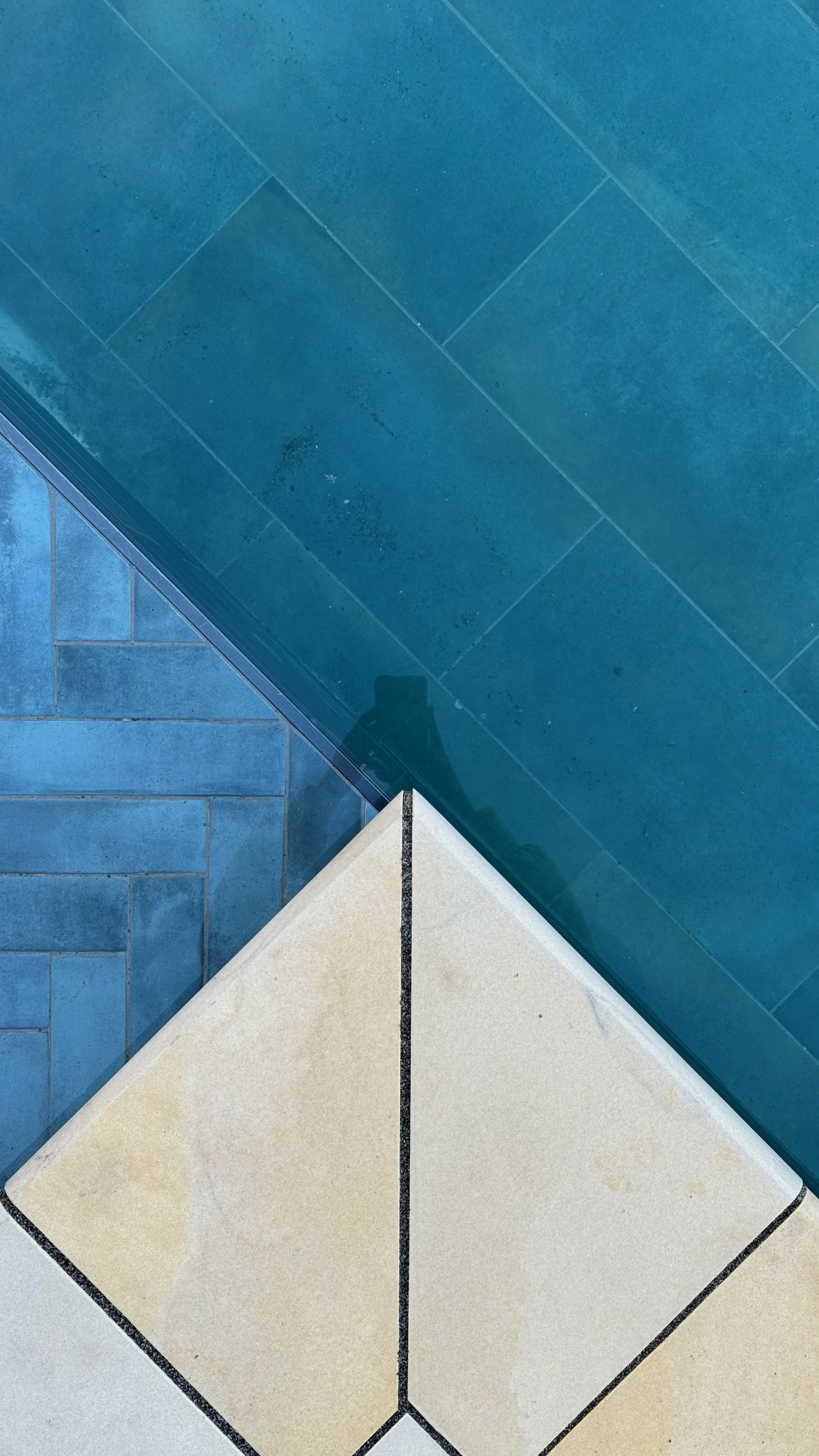 Close-up of a corner where blue, beige, and transparent glass tiles intersect, with a visible reflection of a person taking the photo.