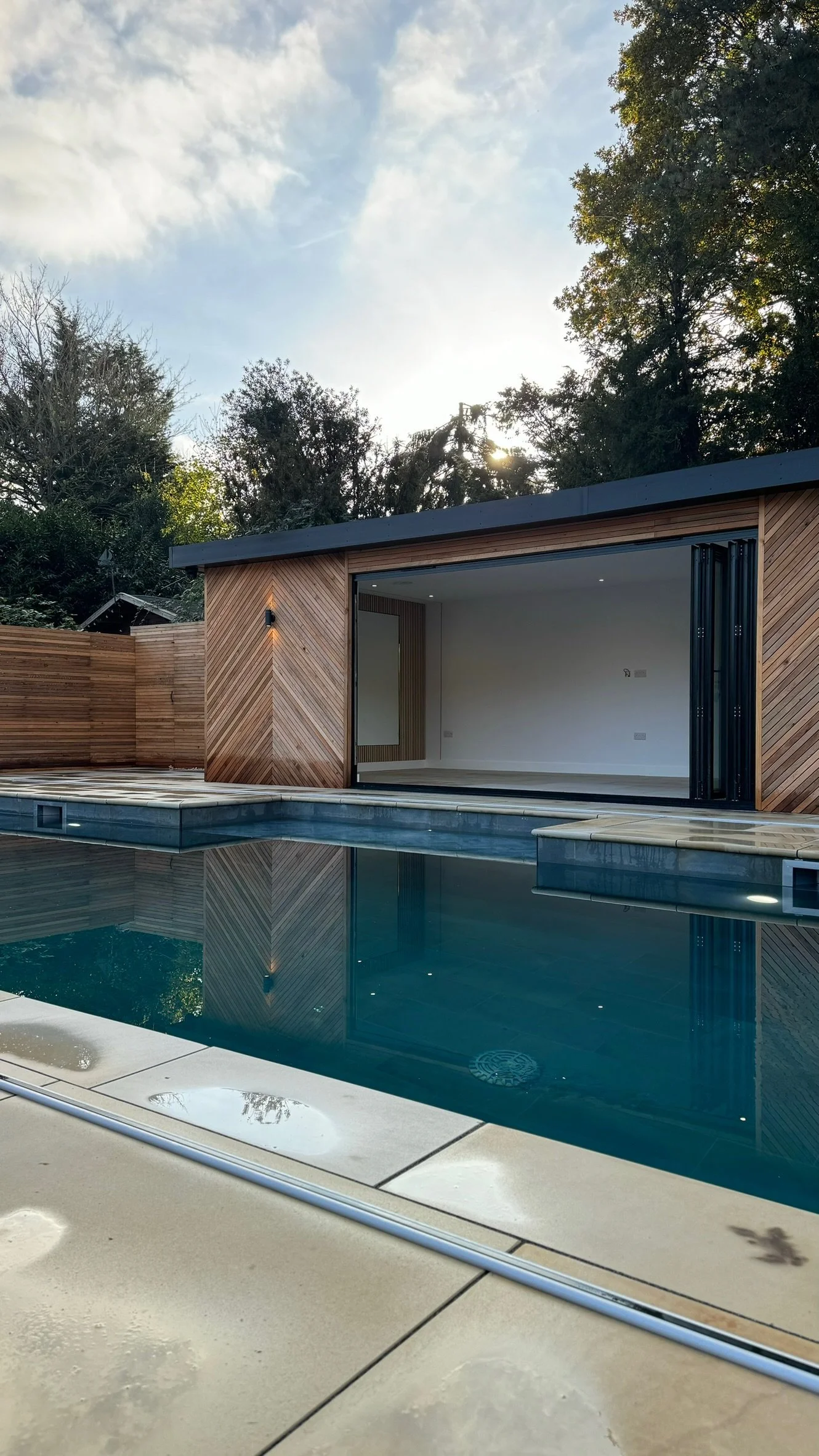 Modern house with a wooden exterior, large sliding glass doors, and a swimming pool in the foreground, surrounded by trees and a partly cloudy sky.