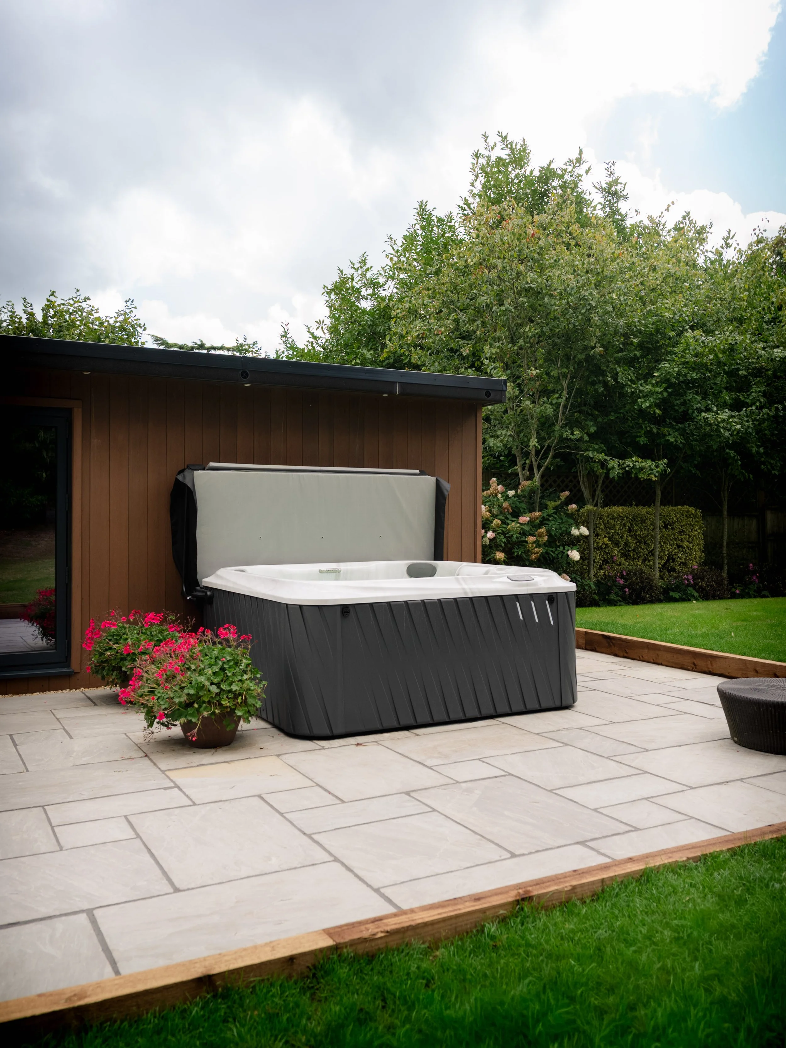 Outdoor hot tub on a stone patio next to a brown house with a wooden border. There is a pink flowering plant in a pot beside the hot tub, and a grassy yard with trees and bushes in the background.