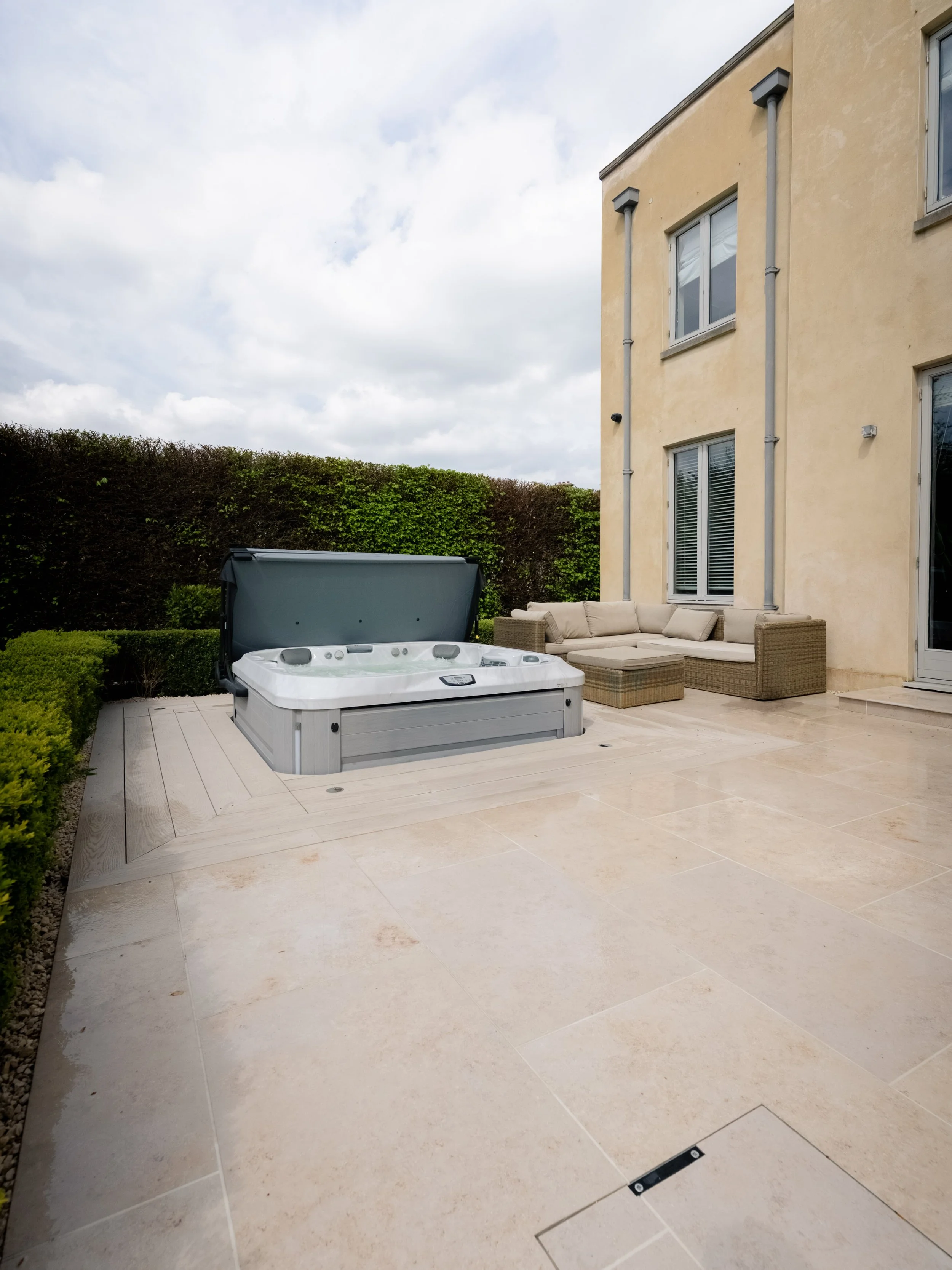 Outdoor patio with a hot tub and a beige L-shaped outdoor sofa set next to a multi-story beige house, against a backdrop of a green hedge and a cloudy sky.