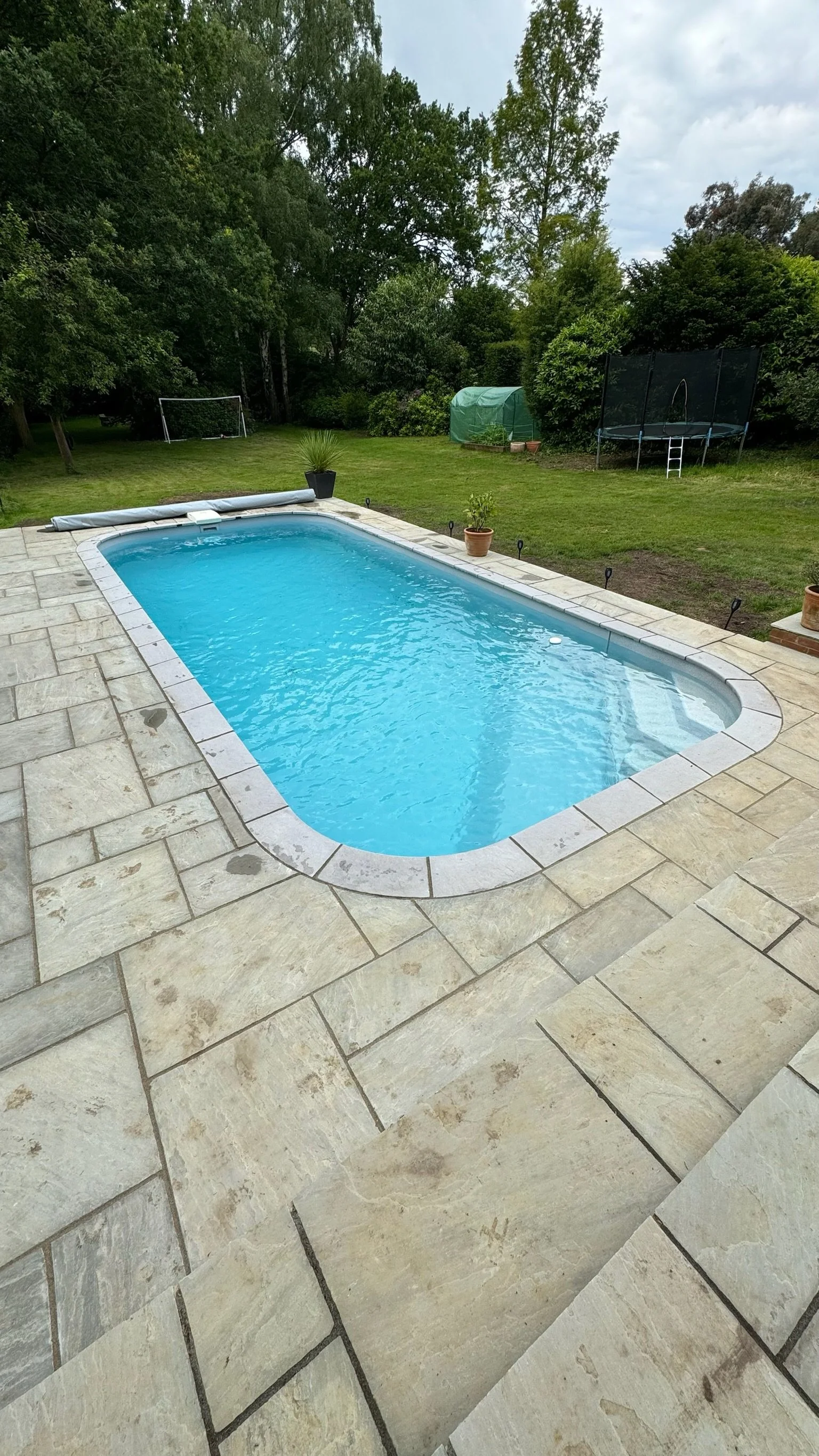 A rectangular swimming pool with light-colored tile surround in a backyard, bordered by a grassy yard with trees, a trampoline, and a small greenhouse.