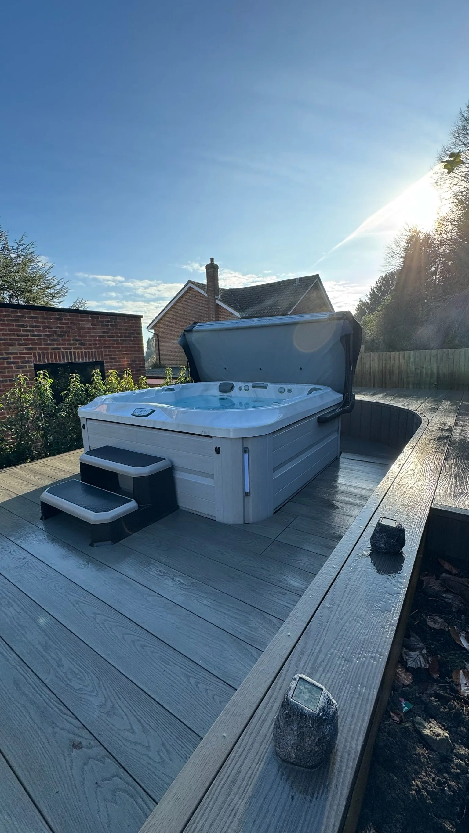 Outdoor hot tub on a wooden deck with steps, in a backyard under a blue sky with sunlight.