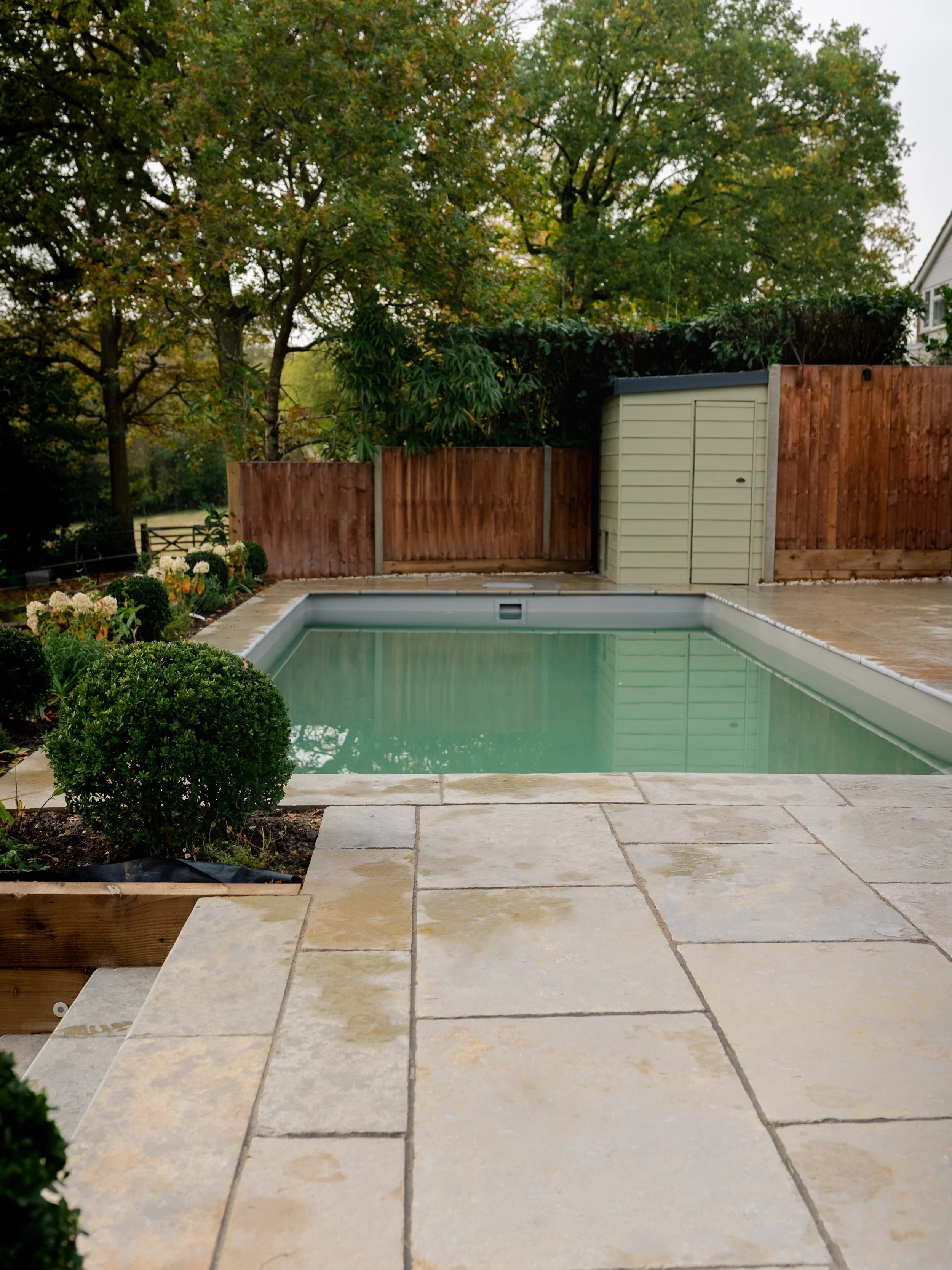 A backyard swimming pool surrounded by stone patio and landscaped bushes, with a wooden fence and trees in the background.
