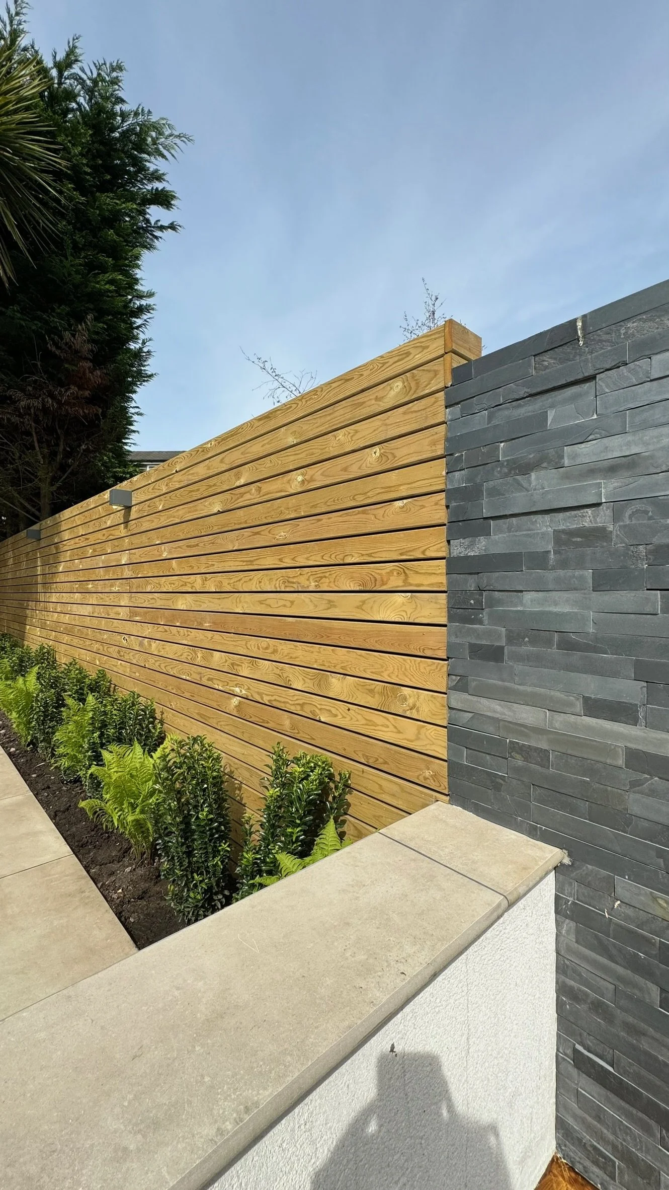 A modern privacy fence with horizontal wooden slats and a black stone wall section, accompanied by a flower bed with green shrubs, in a sunny outdoor setting. Esme are a widely trusted garden design and landscaping company serving surrey.