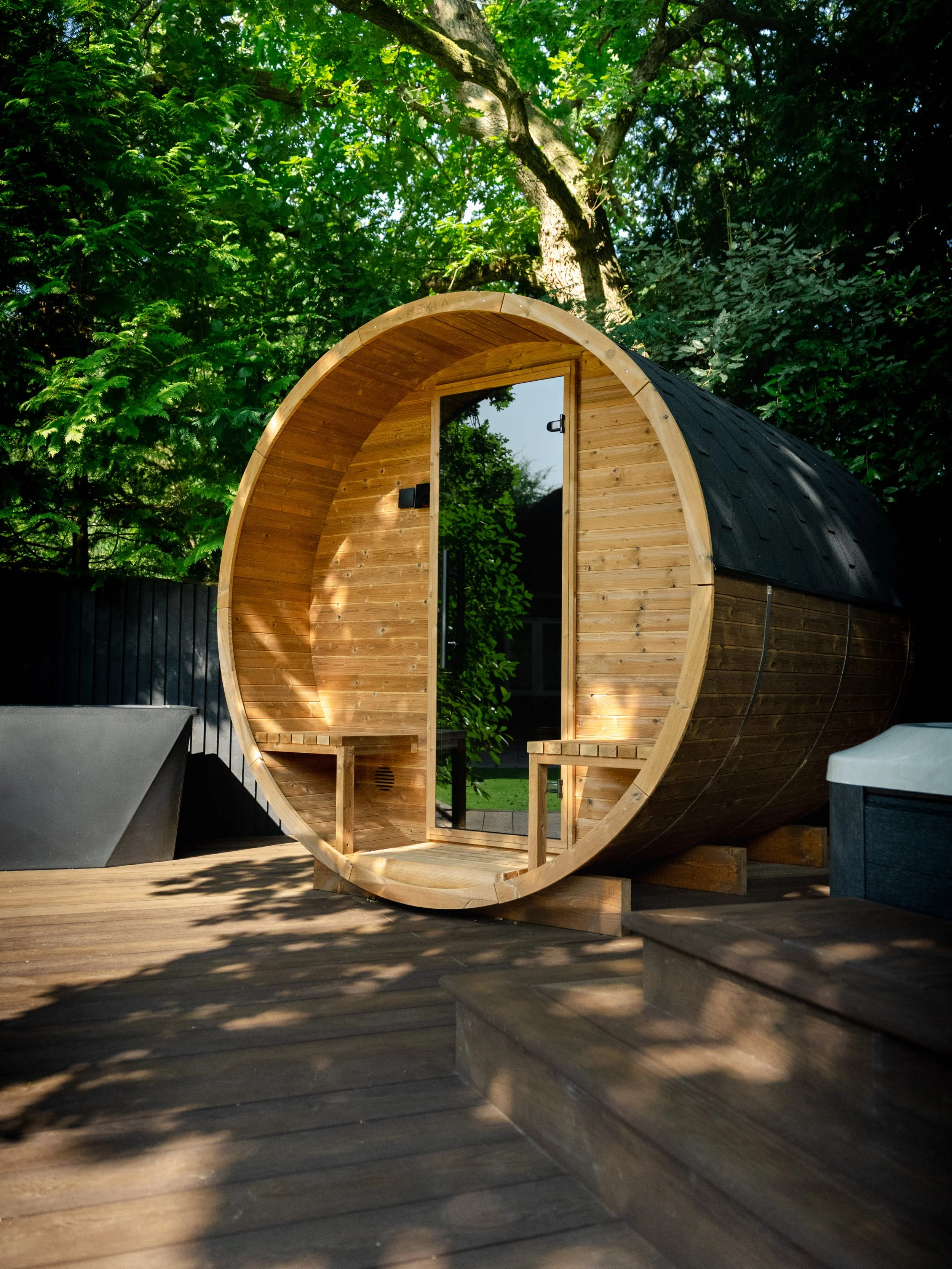 A wooden barrel-shaped sauna is outdoors among trees and greenery, with a glass door and benches inside. Expert landscapers and garden designers in Surrey, Hampshire, Berkshire, Camberley, London and surrounding areas.