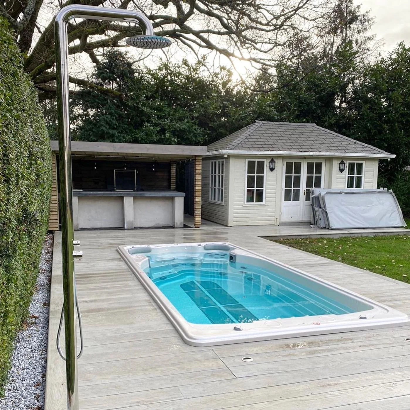 Backyard with a hot tub, outdoor shower, small house, and an outdoor kitchen area with a sink and chimney, surrounded by trees and a green lawn.