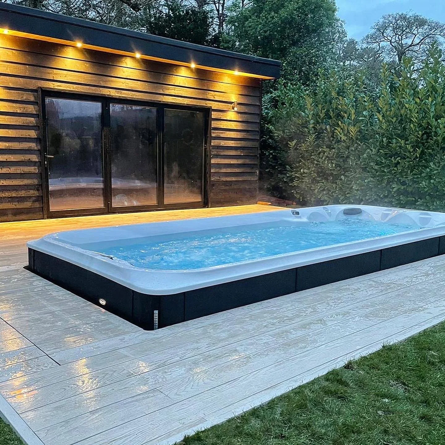 Outdoor hot tub on a white deck with a wooden house and tall green bushes in the background during dusk.