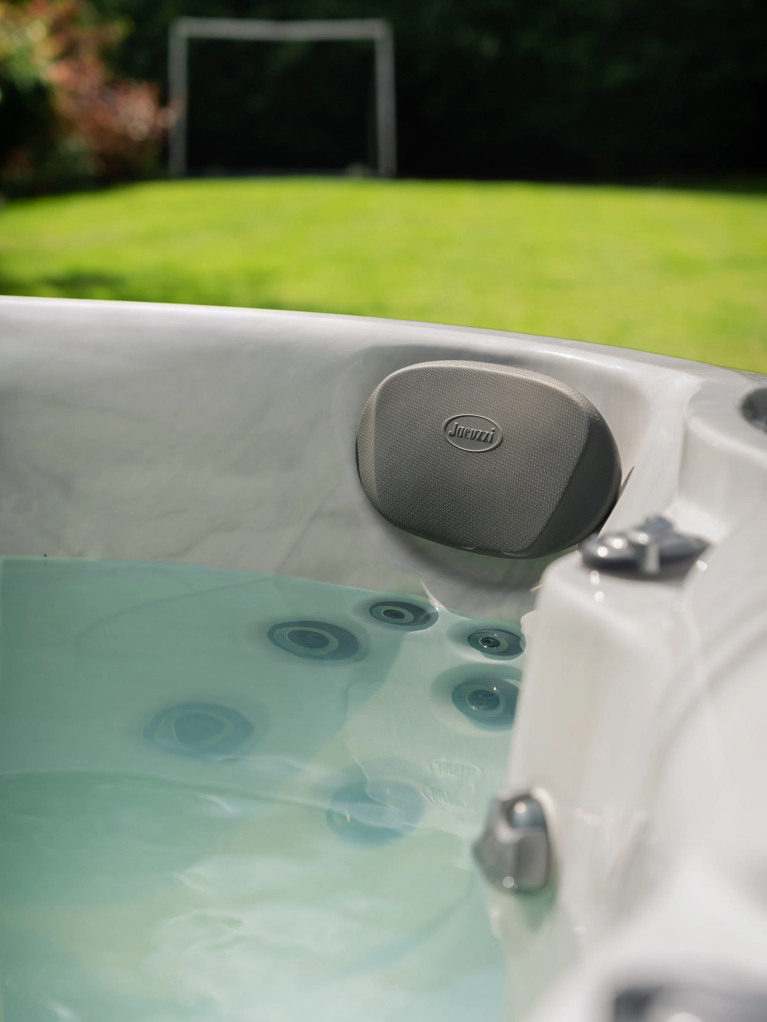Close-up of a jacuzzi hot tub with water and speakers in an outdoor backyard scene with a grass lawn and soccer goal in the background.