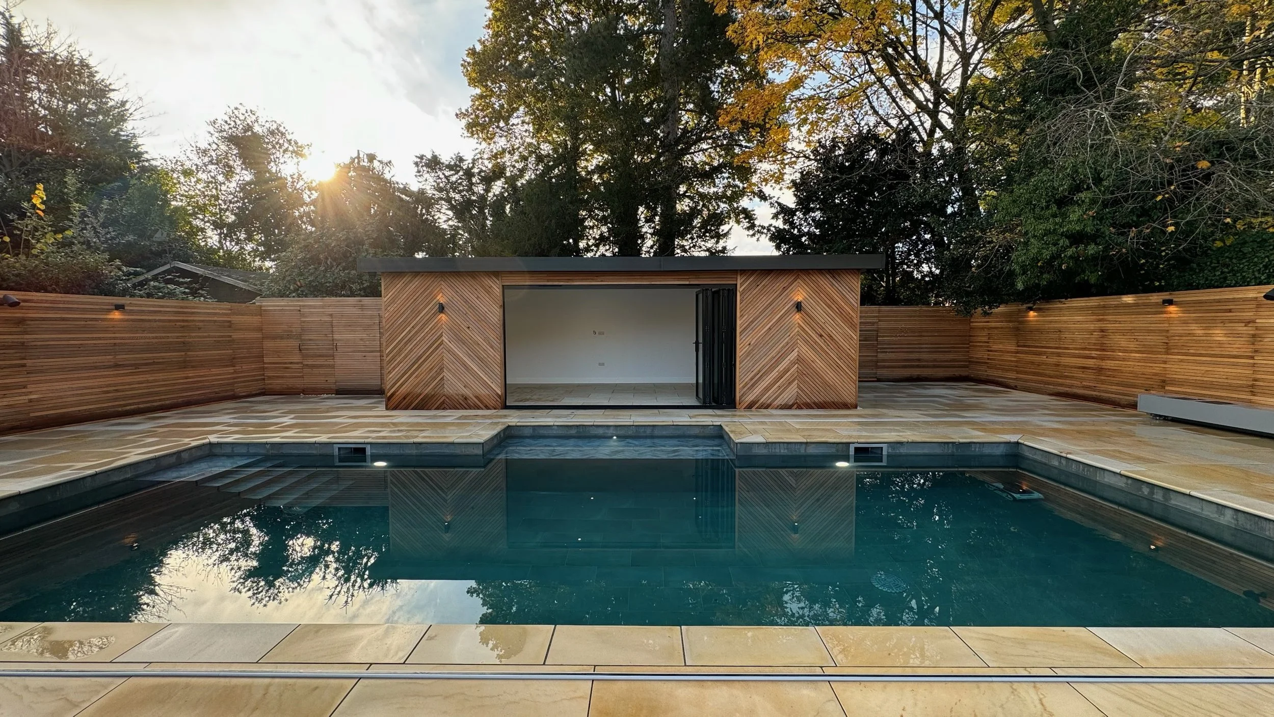 A backyard with a swimming pool and a modern pool house with wooden walls and black sliding doors, surrounded by a wooden privacy fence and greenery, during sunset.