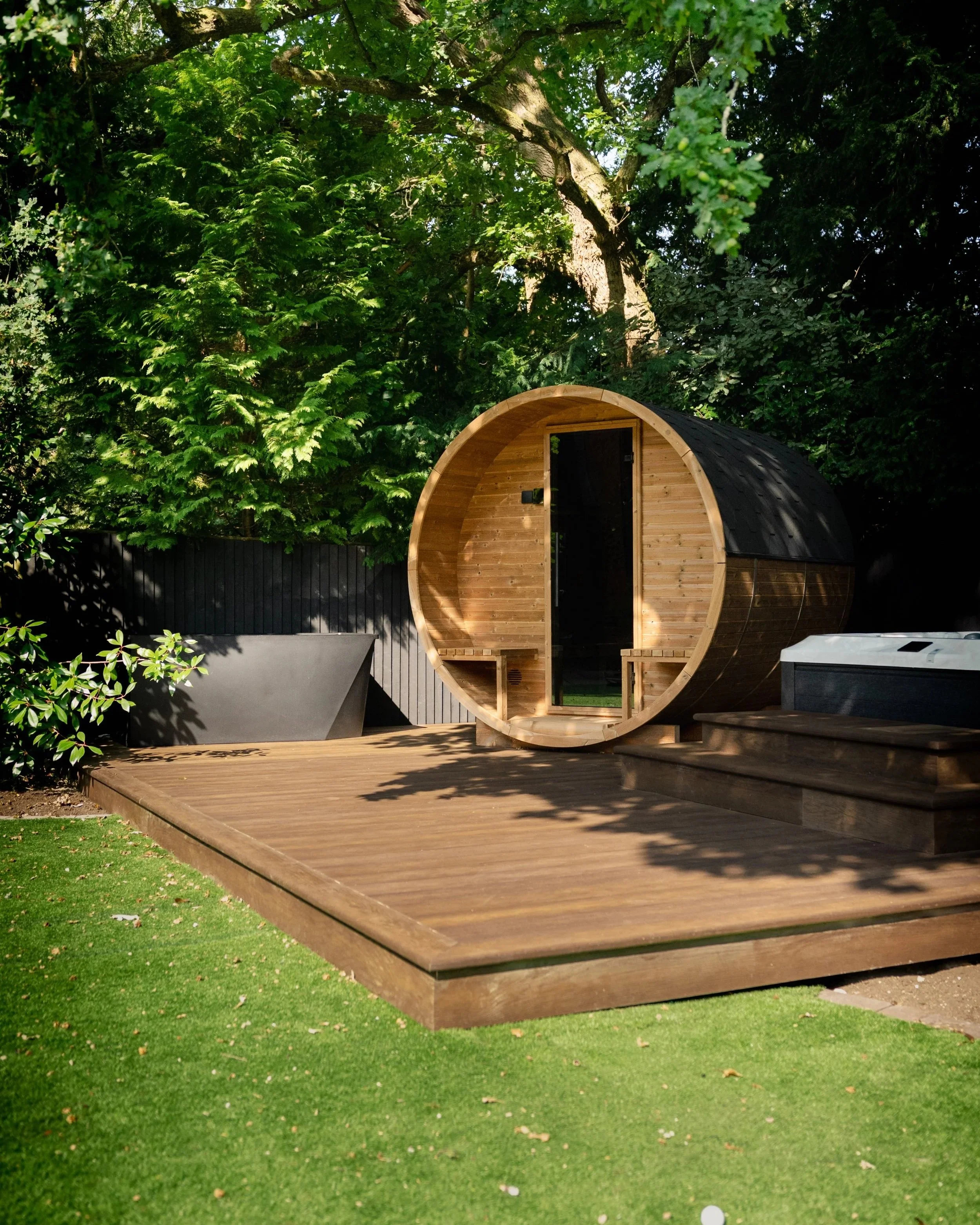 A wooden barrel-shaped sauna on a wooden deck surrounded by green trees and grass. Expert landscapers and garden designers in Surrey, Hampshire, Berkshire, Camberley, London and surrounding areas.
