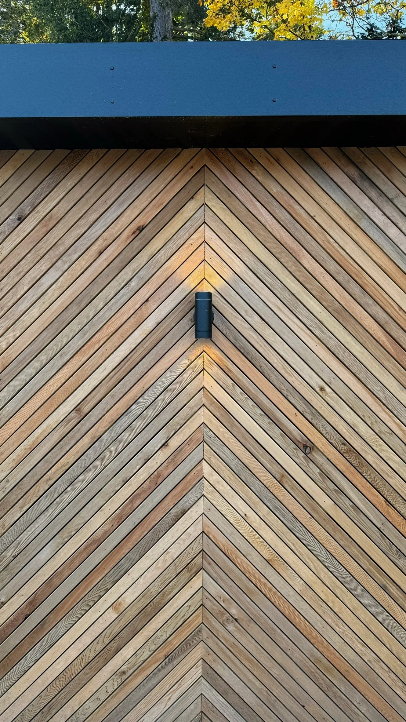 Close-up of a building exterior with diagonal wooden planks forming a V-shape, a black cylindrical outdoor light fixture at the center, and a black accent overhang at the top.