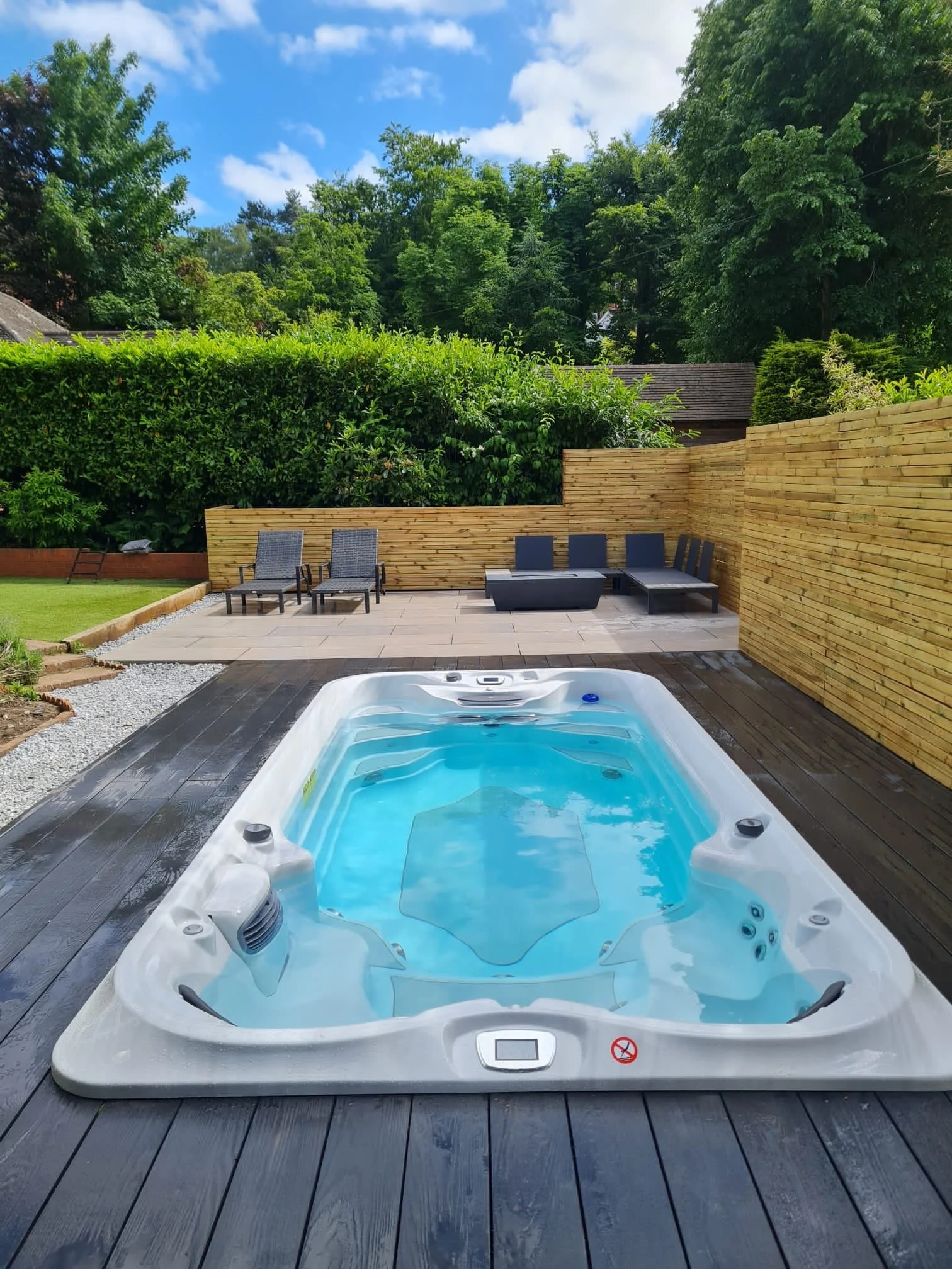 Backyard with hot tub on wooden deck, lounge chairs, and a privacy fence, surrounded by green trees and a blue sky with clouds.