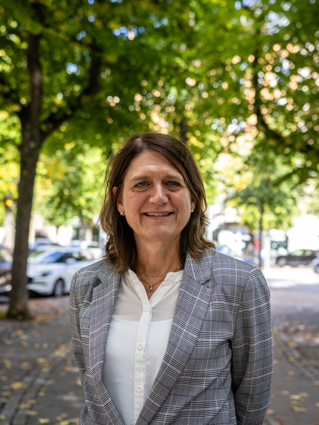 A woman in a plaid blazer and white top smiling outdoors under green trees with parked cars in the background.
