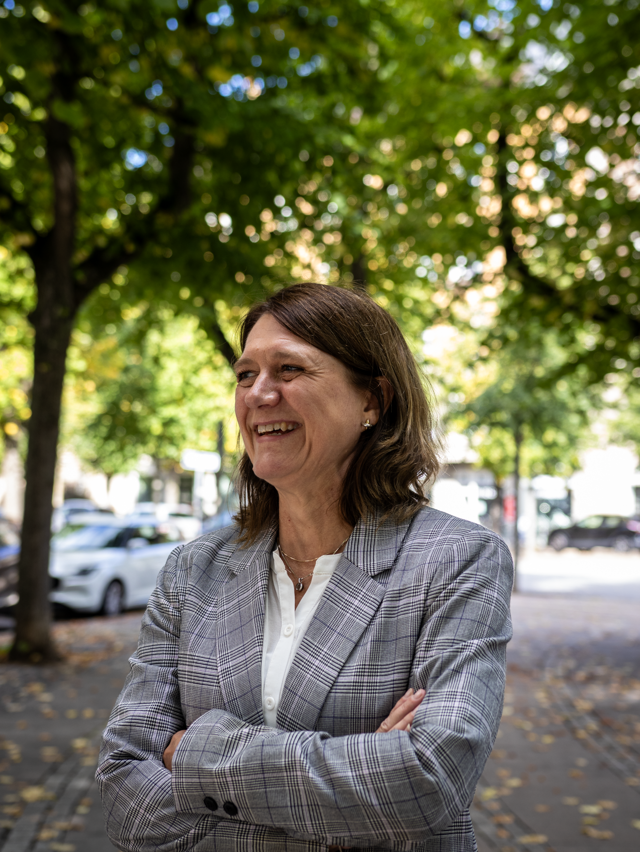 A woman with shoulder-length brown hair smiling and standing outdoors with her arms crossed, wearing a gray plaid blazer and a white shirt, in front of green-leaved trees and parked cars.