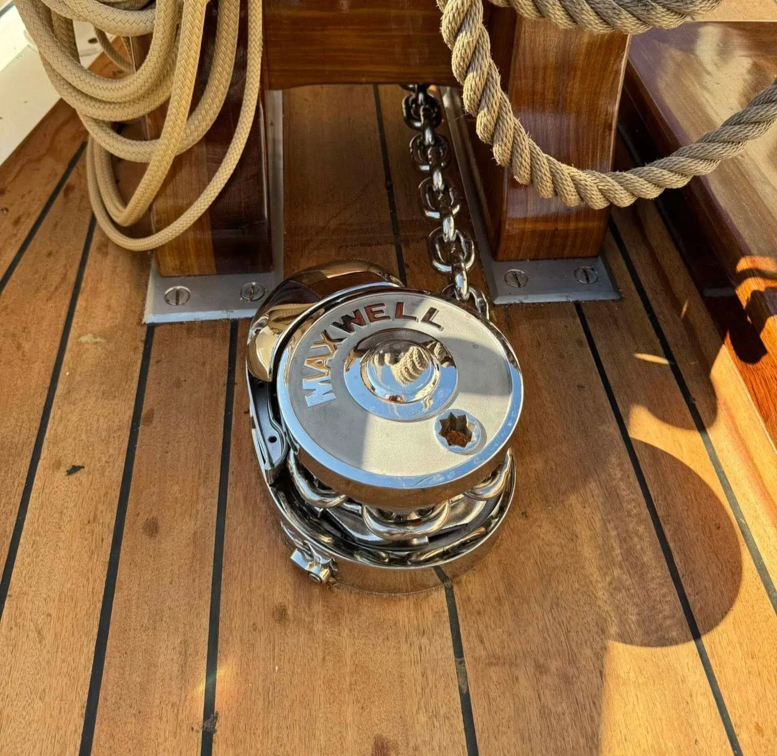 Metal ship steering wheel with the brand name 'Maywell' on top, attached to the wooden deck of a boat, with ropes and hardware visible nearby.