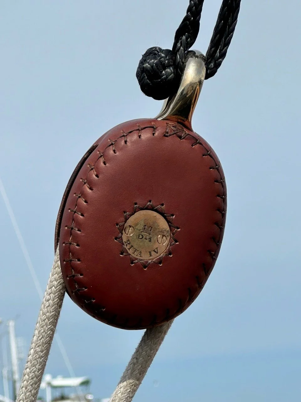 Close-up of a brown leather pulley with a metal center, hanging from black ropes on a boat, against a cloudy sky.