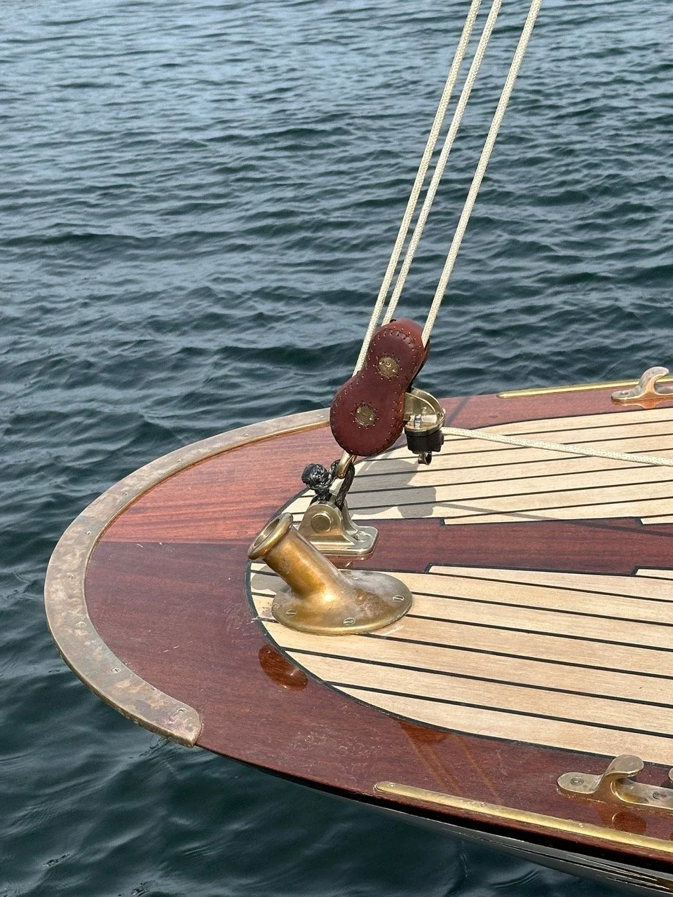 Close-up of a wooden yacht bow with brass cleat, part of the deck, and water in the background.