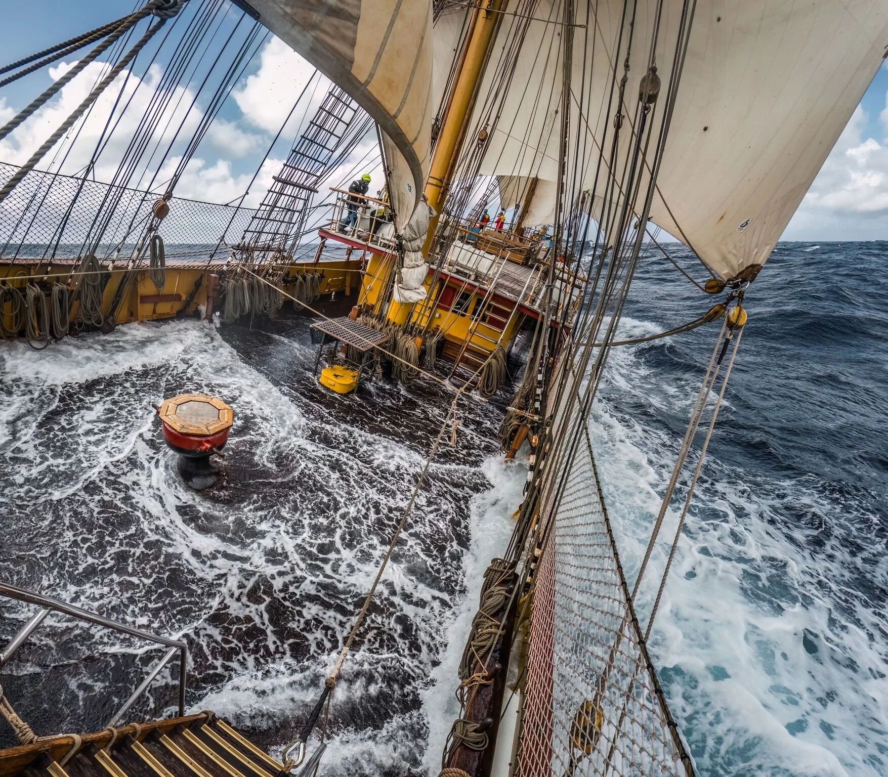 View from a sailing ship's bow looking towards the deck with sailors, rigging, and sails in open ocean under cloudy sky.