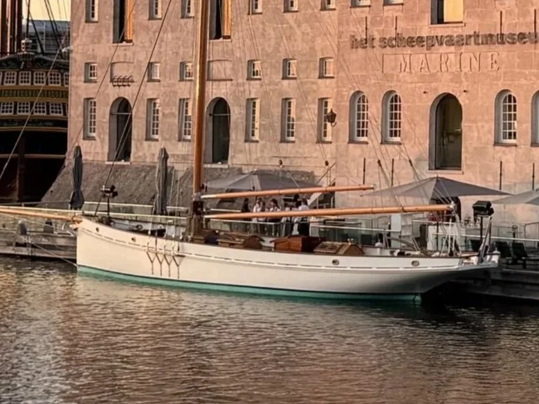 A white sailboat with a wooden mast docked by a stone building with large windows and a sign reading 'de scheepvaartmuseum MARINE' in the background.