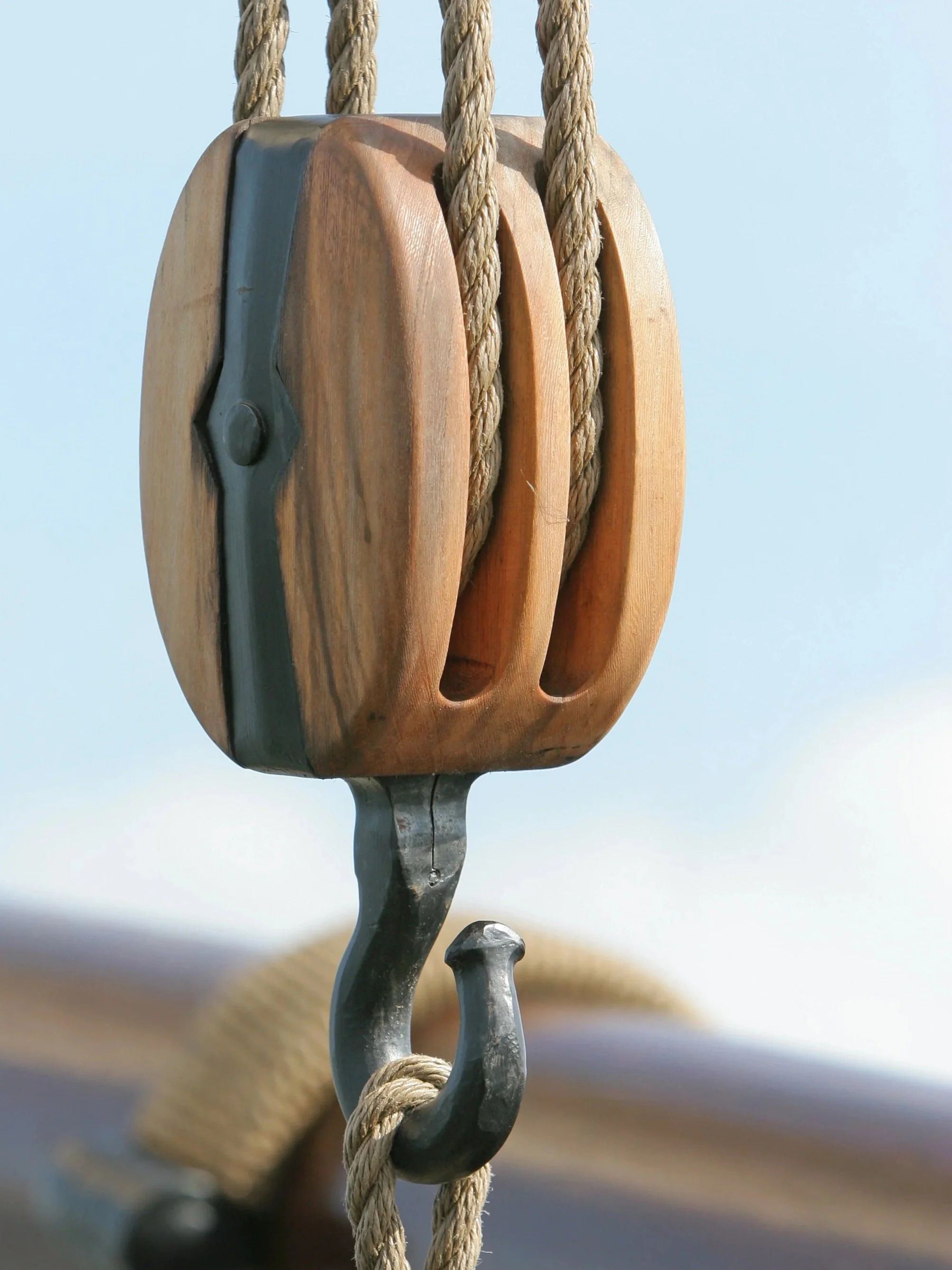 Close-up of a vintage pulley with a wooden outer shell, connected to thick, beige rope, against a light blue sky.