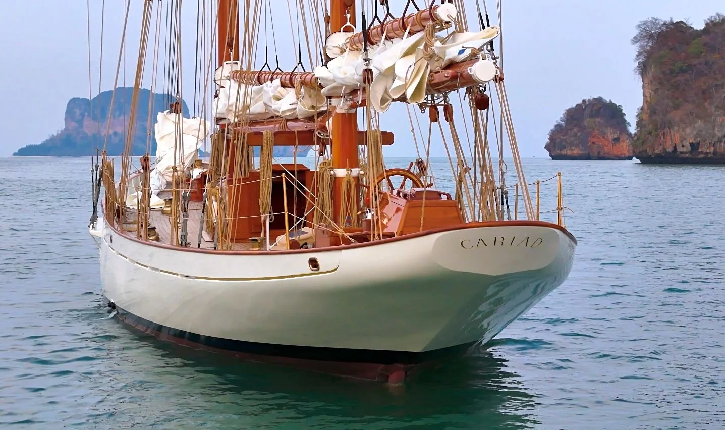 A wooden sailing yacht named Cariad with its sails furled, sailing in calm waters near rocky islands.