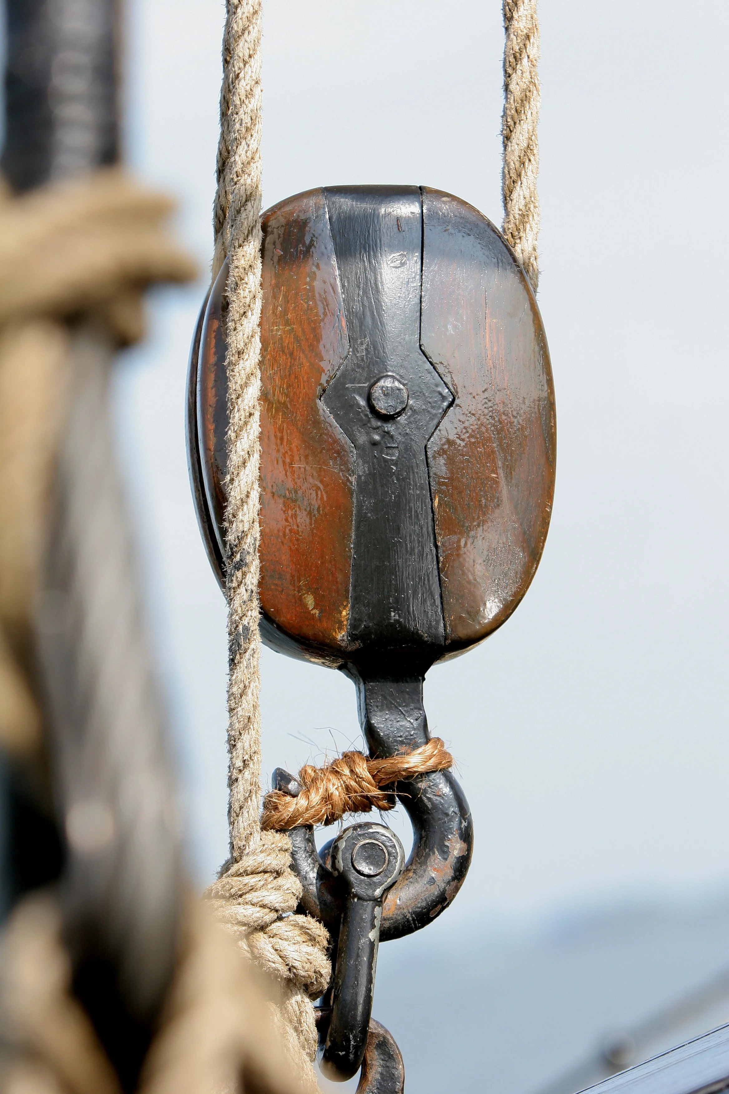 Close-up of a metal pulley with a black hook and rope, mounted on a boat or ship, with a cloudy sky in the background.