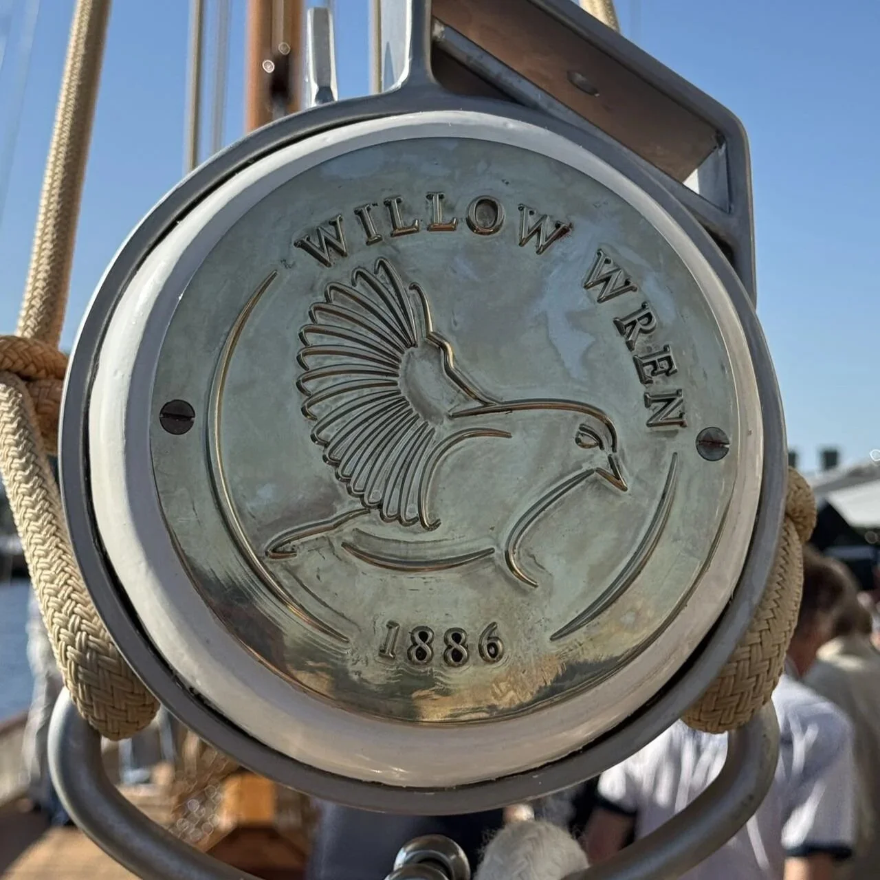 Close-up of a circular metal sign with a bird and the text 'Willow Wren 1886' on it, being held by a person with ropes and a boat in the background.