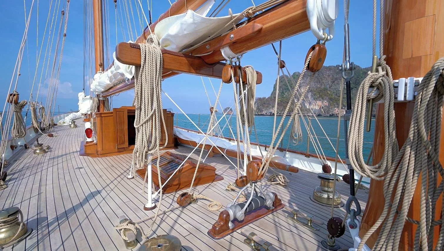 View of a sailboat's deck with ropes, pulleys, and wooden fittings, overlooking a coastline with rocky islands and blue sky.