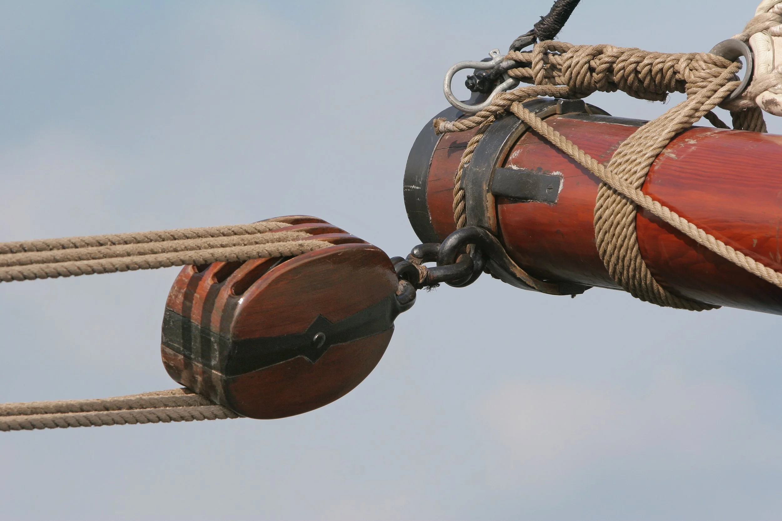 Close-up of rigging on a tall ship, showing wooden pulley blocks and thick ropes against a cloudy sky.