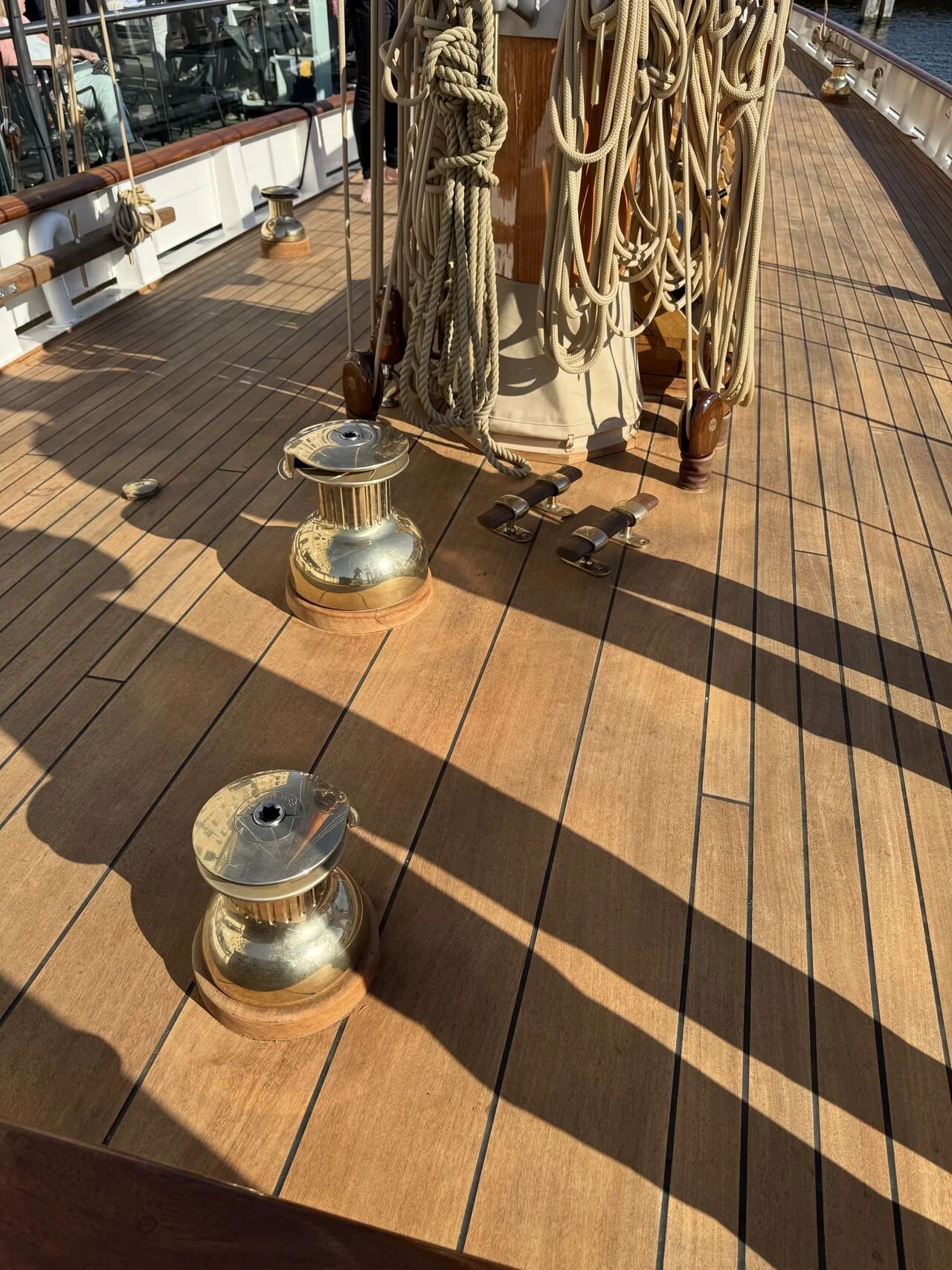 Deck on a ship with brass cleats, coiled ropes, and a winch, with shadows cast on the wooden surface.