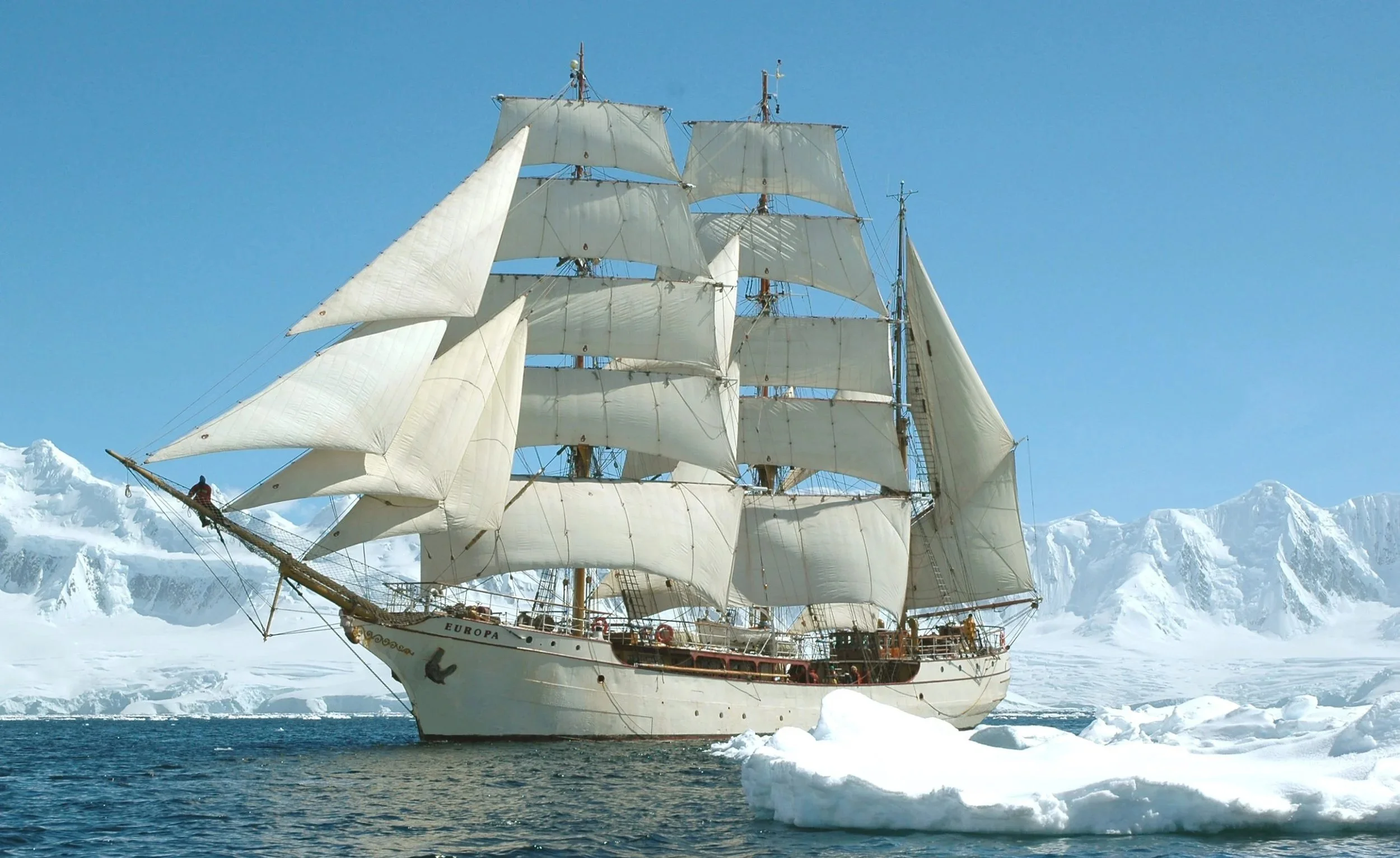 A tall sailing ship with white sails navigating through icy waters with snow-covered mountains in the background.