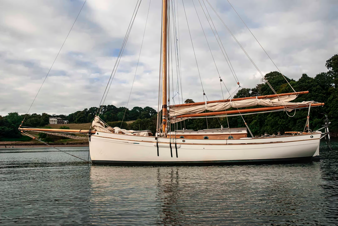 A white sailboat with a single mast floating on calm water with a wooded shoreline and a building in the background, under a cloudy sky.