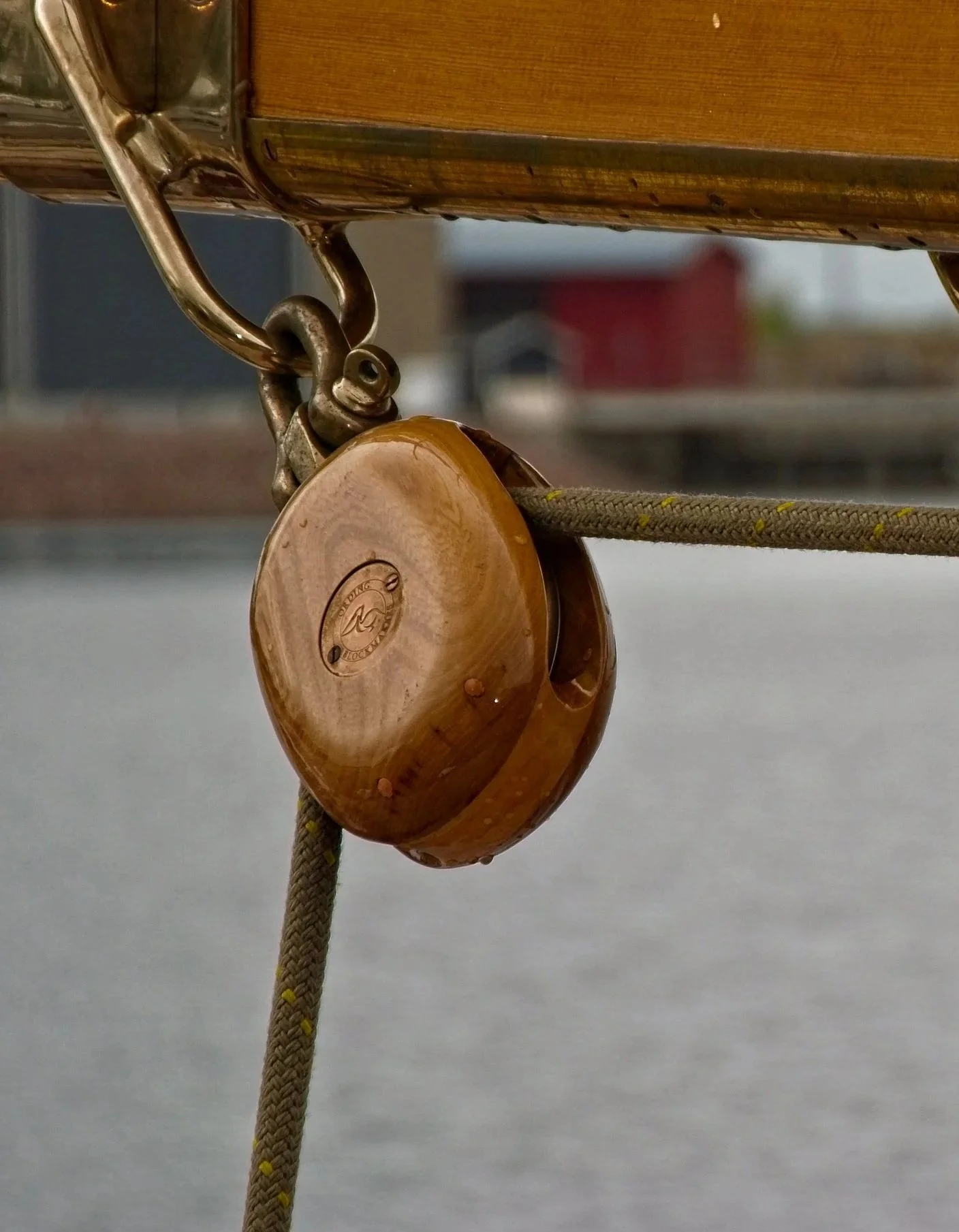 Close-up of a round, wooden pulley attached to a rope, part of a boat's rigging, with water and a distant shoreline in the background.