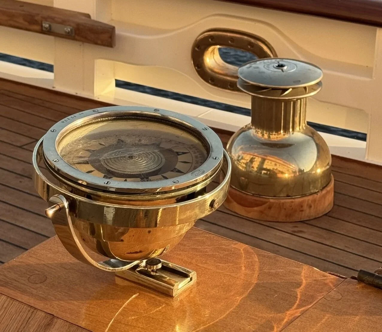 A brass navigation compass and a brass night lantern on a wooden surface outside on a boat deck.