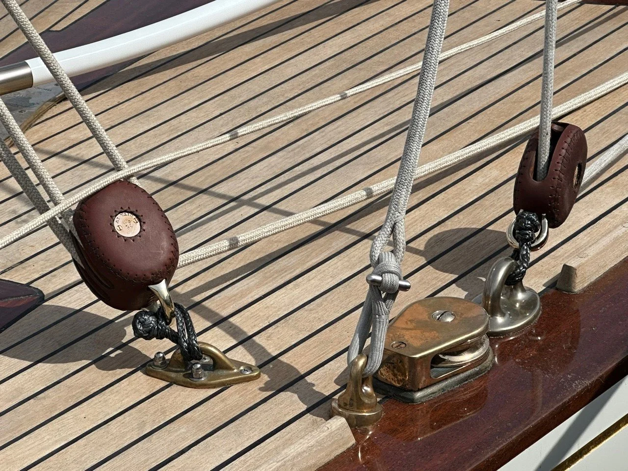 Close-up of a yacht's deck hardware, showing metal fittings, ropes, and leather-coated turnbuckles on a wooden surface.