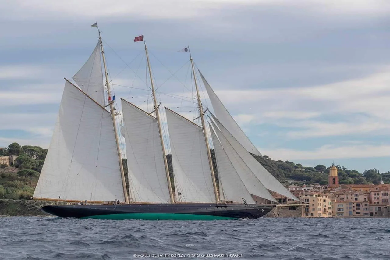 A large sailing ship with white sails glides past a coastal town with buildings and a tower in the background.