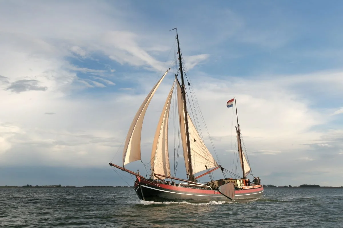 A sailboat with beige sails sailing on the water under a partly cloudy sky with a Dutch flag.