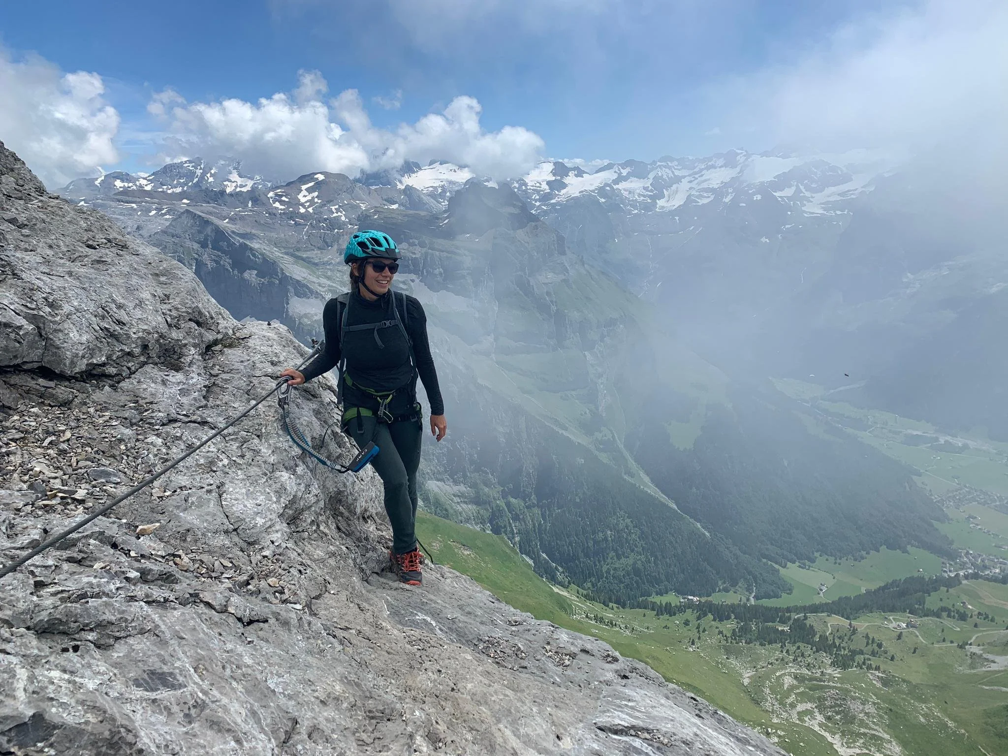 Frau in Kletterausrüstung und Helm klettert an einer Felswand in den Bergen mit Blick auf Tal und schneebedeckte Berge im Hintergrund.