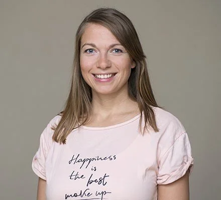 Lächelnde junge Frau trägt ein rosa T-Shirt mit der Aufschrift 'Happiness is the best make-up' auf grauem Hintergrund.