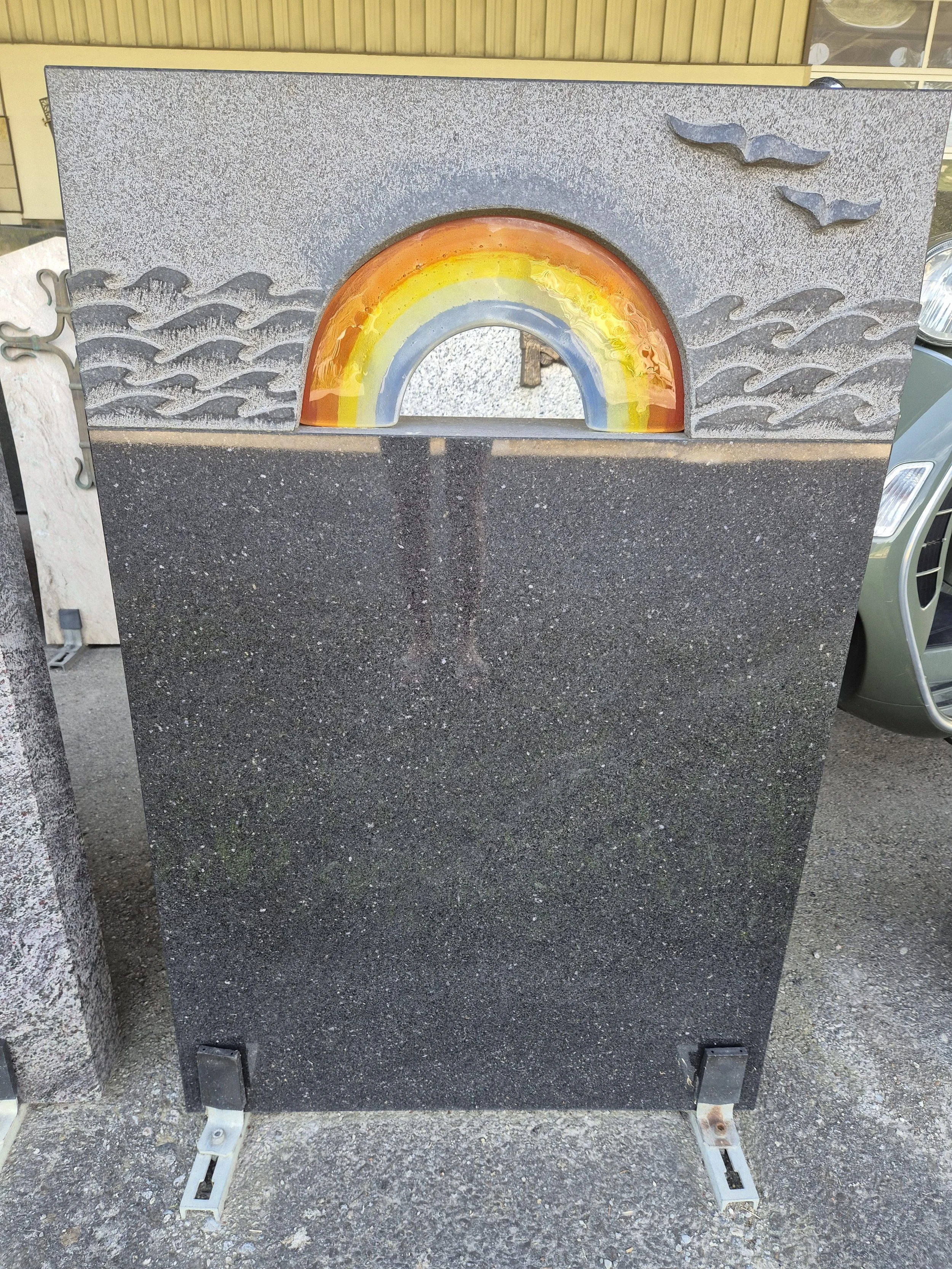 Ein Wandbild mit einem Regenbogen und Flugvögeln. Der Regenbogen ist bogenförmig mit Farben in Orange, Gelb, Blau und Grau, eingebettet in eine graue Wand mit Wellendesign. Es ist im Freien aufgestellt.