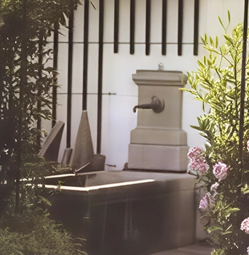 Gartenbrunnen mit Wasserhahn, umgeben von Pflanzen und an einer Mauer mit Gitterstäben.