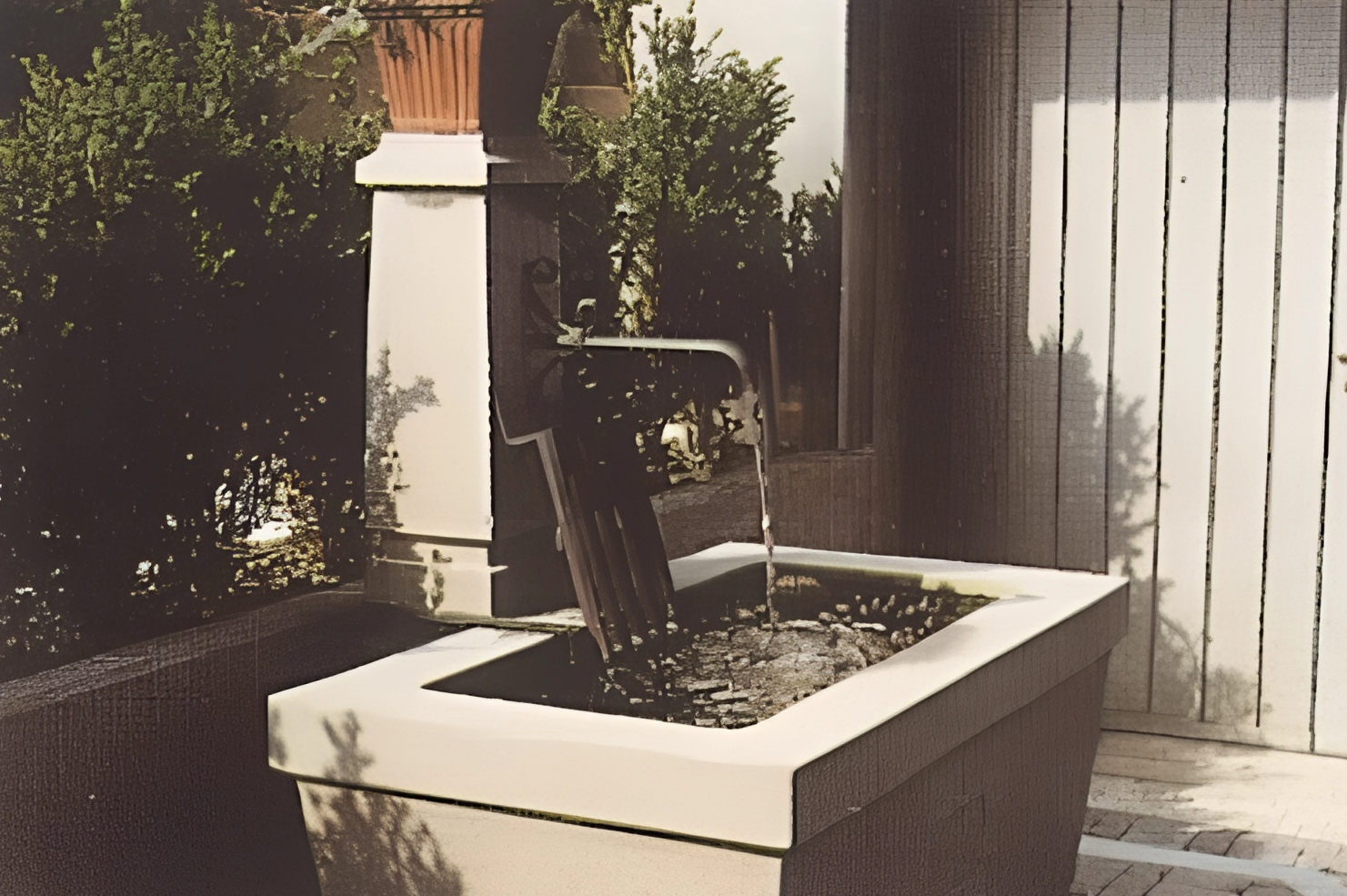 Wasserspiel in einem quadratischen Pflanzkübel mit einem Holzstab, der Wasser nach oben sprudelt, umgeben von Pflanzen und einer Holzlatwando