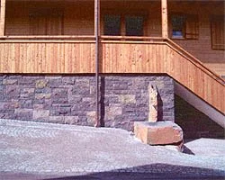Ansicht eines Hauses mit Steinmauer und Holzverkleidung, Treppen und einem Steinblock im Vordergrund.