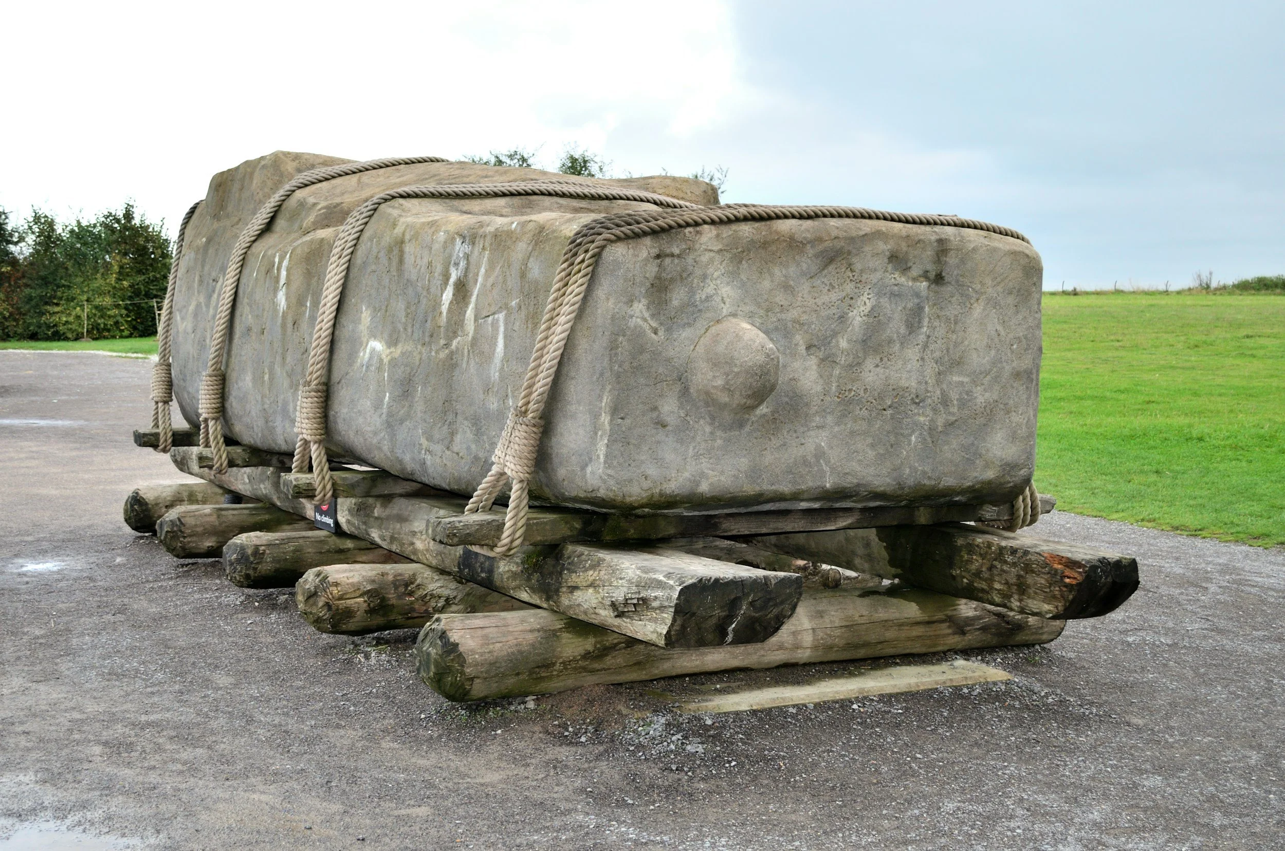 Großer Steinblock auf Holzschlitten, mit Seile befestigt, im Freien auf einer gepflasterten Fläche, im Hintergrund Bäume und eine grüne Wiese.