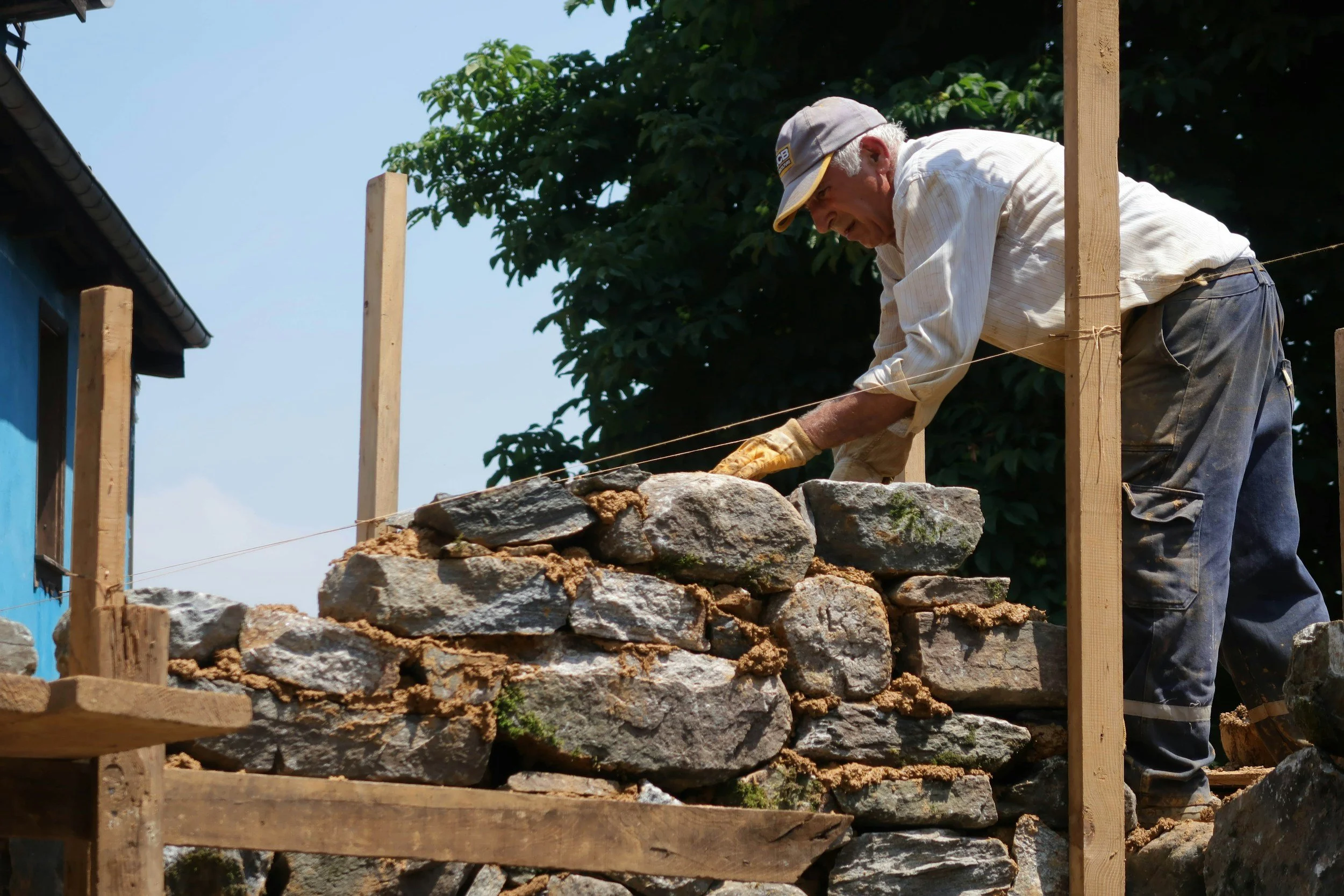 Ein älterer Mann baut eine Trockenmauer aus Steinen auf einer Baustelle im Freien, umgeben von Holzständern.