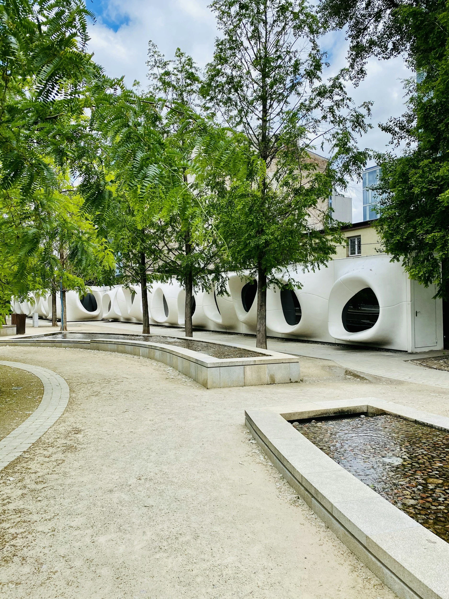 Ein moderner Park mit Bäumen, Sandweg und Wasserbecken, im Hintergrund eine weiße Gebäude mit runden Fenstern.